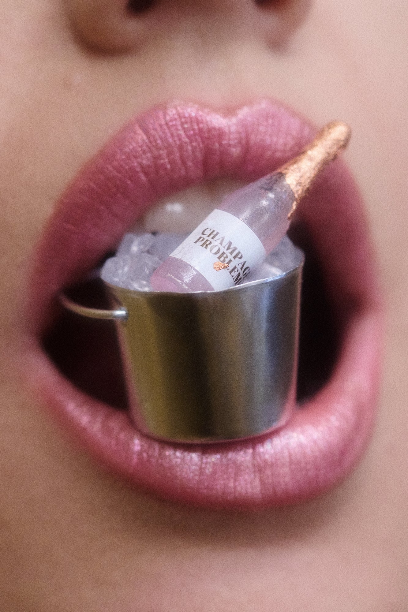A close-up portrait of a person's lips holding a miniature champagne bucket filled with ice and a small bottle.