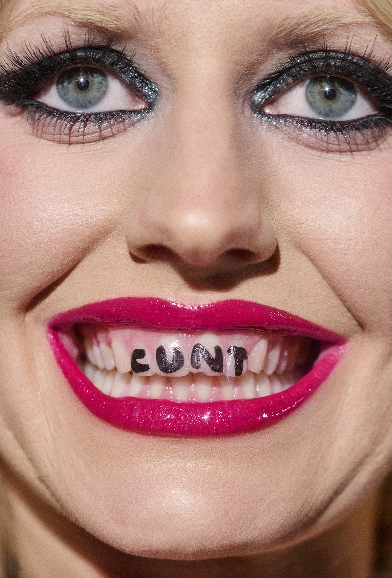 A close-up portrait of a person with bold makeup and text written on their teeth.