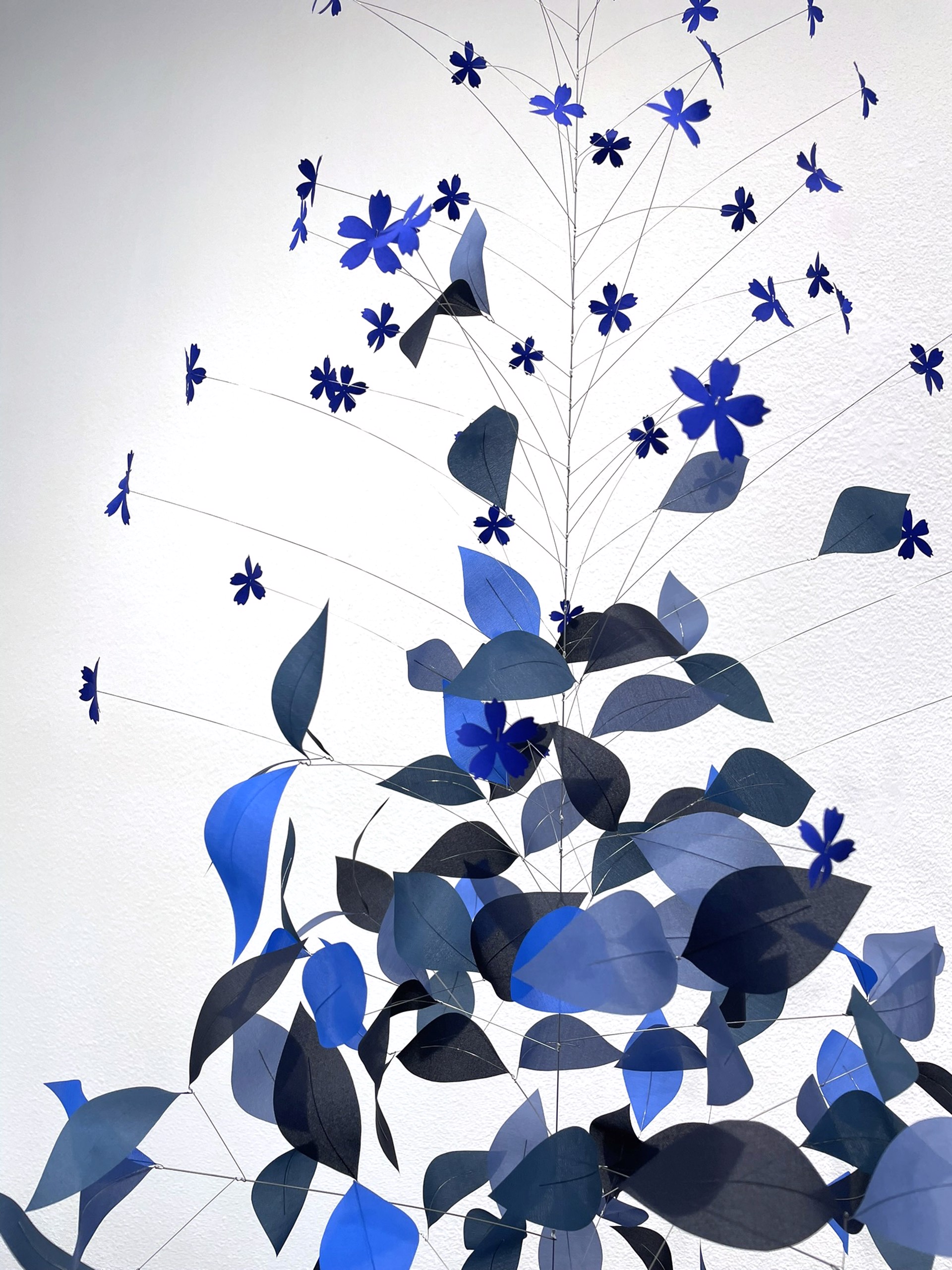 A vertical mobile sculpture featuring cascading blue leaves and star-shaped cutouts against a stark white background.