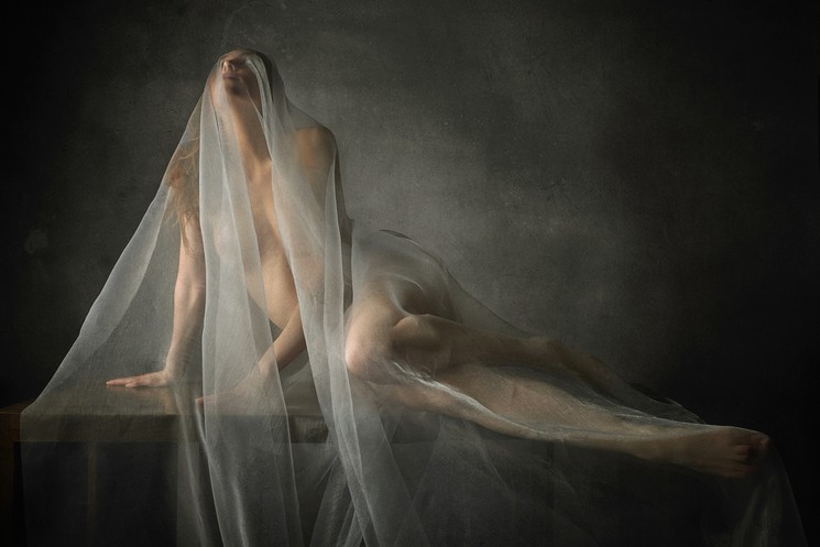 A nude woman with a veil over her head sitting on a table.