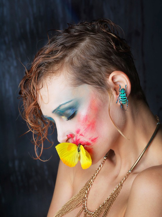 A portrait of a person with vibrant face paint and insect-themed accessories against a dark background.