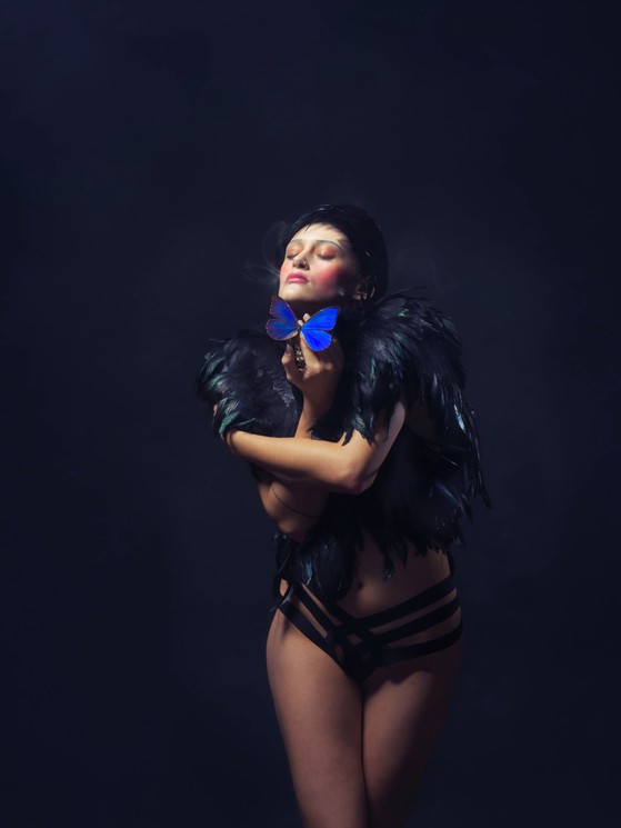 A person in black feathered attire holds a blue butterfly against their neck, eyes closed in a moment of quiet intensity.