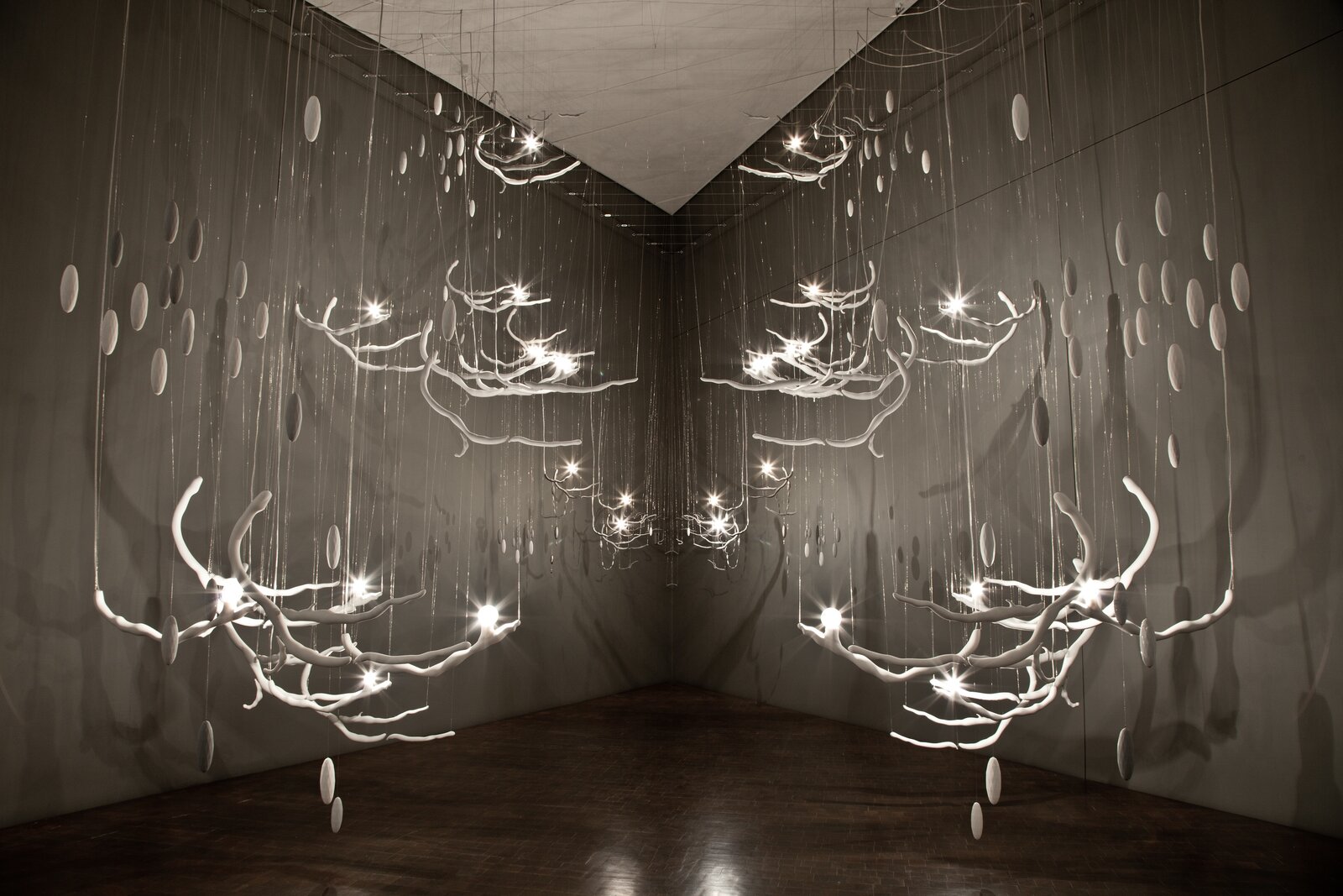 "A suspended installation of white, branch-like structures with glowing lights and floating discs in a monochromatic gallery space."