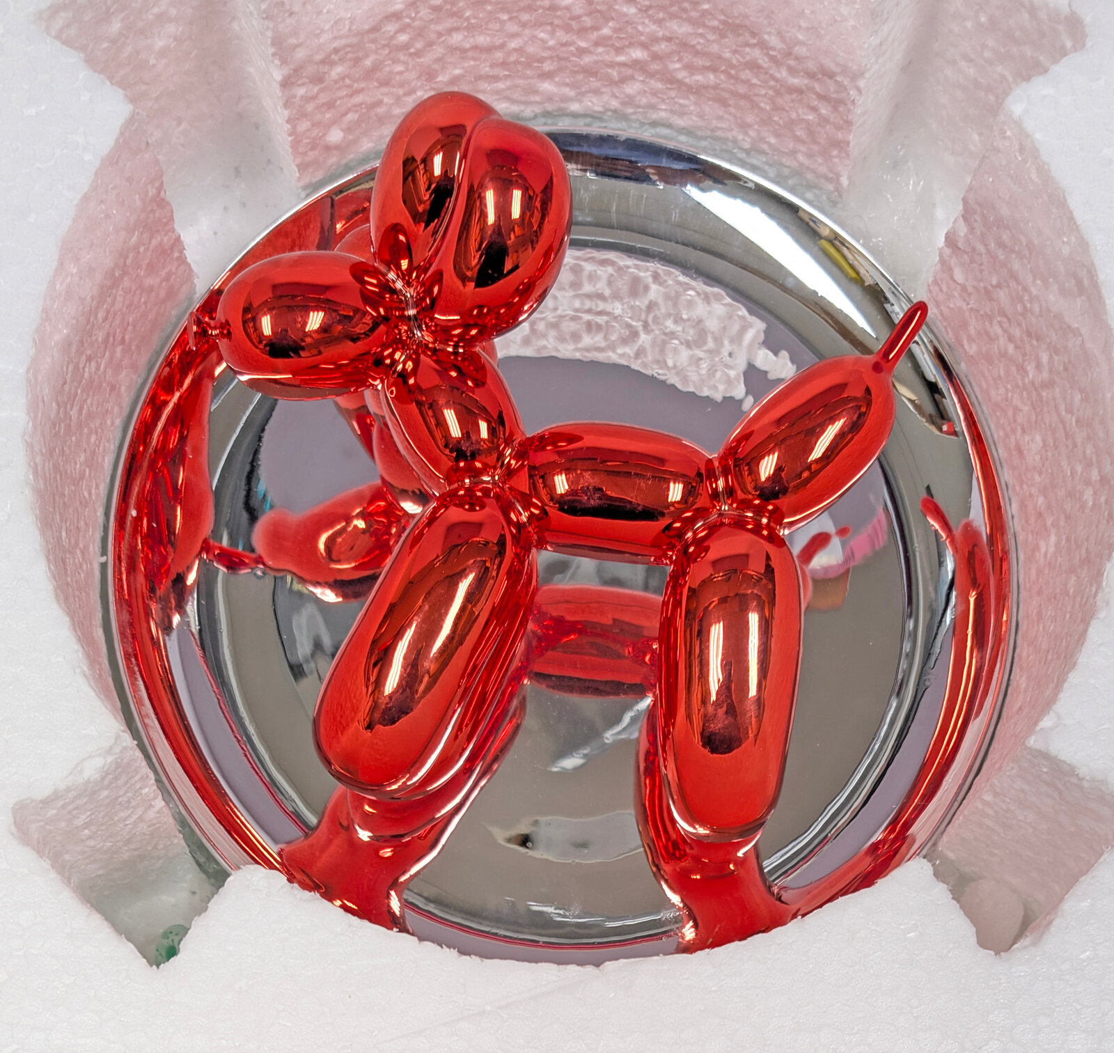 A polished red balloon-dog sculpture displayed within a reflective circular frame.