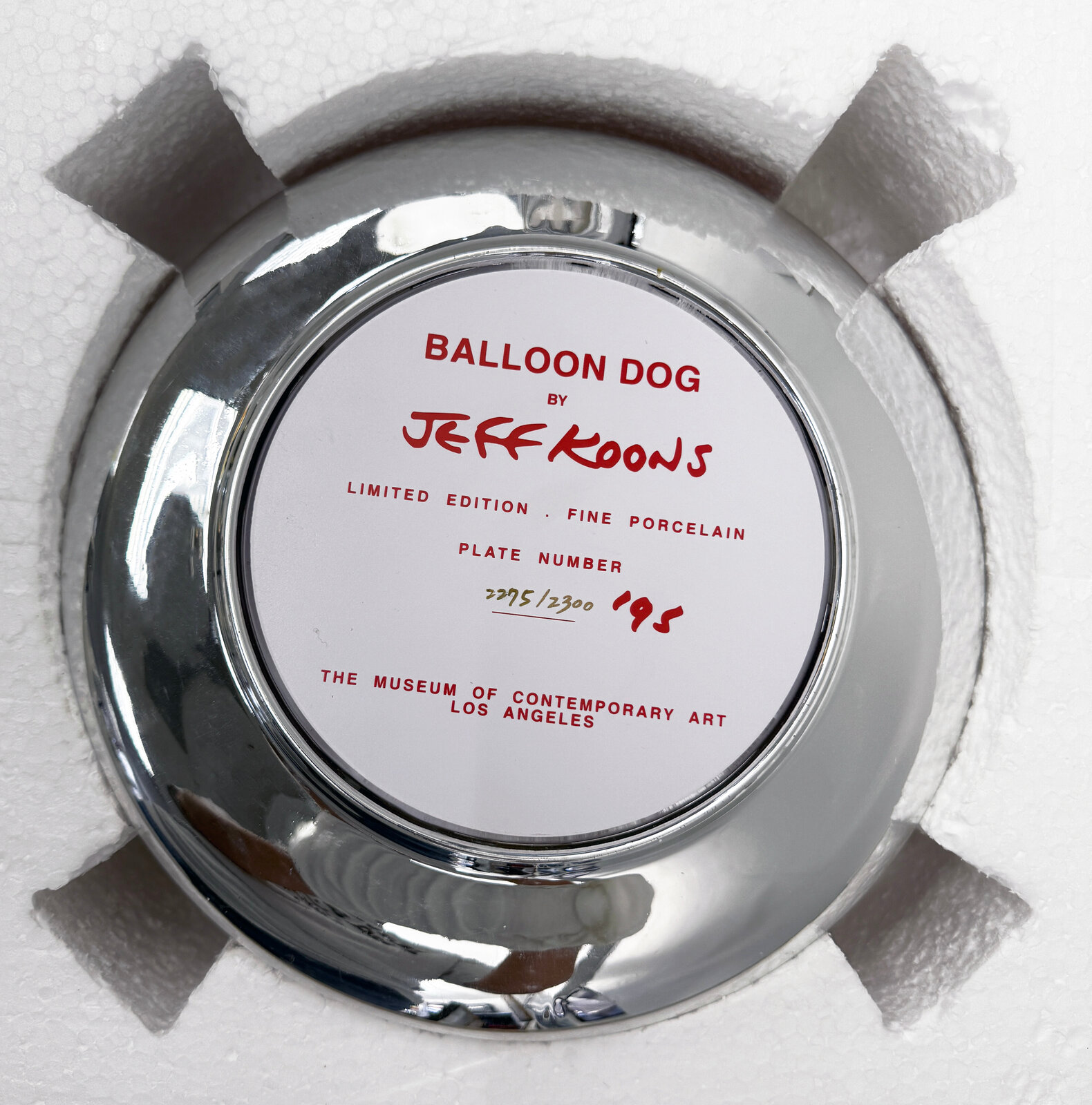 A polished red balloon-dog sculpture displayed within a reflective circular frame.