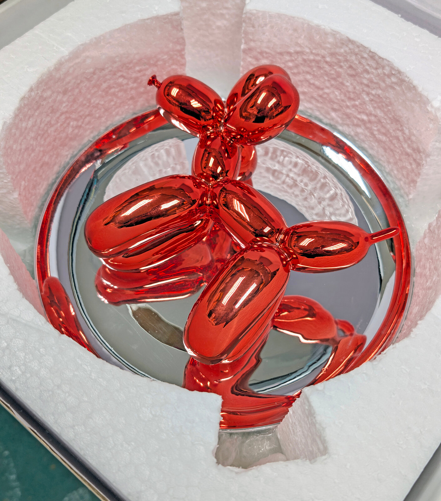 A polished red balloon-dog sculpture displayed within a reflective circular frame.