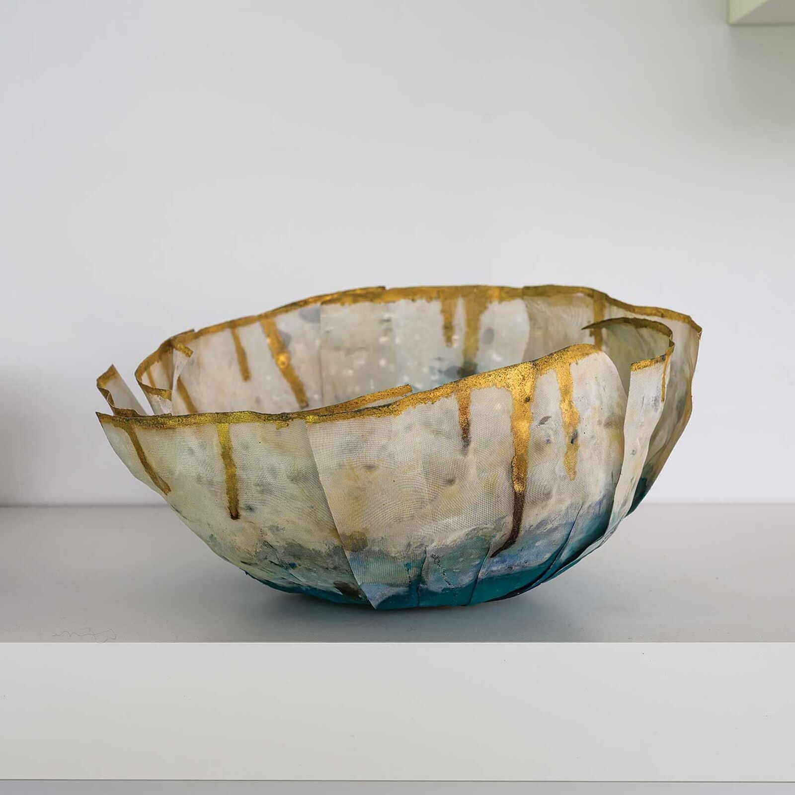 A sculptural bowl with a ruffled, organic form featuring gold leaf drips and a gradient of blue to white.