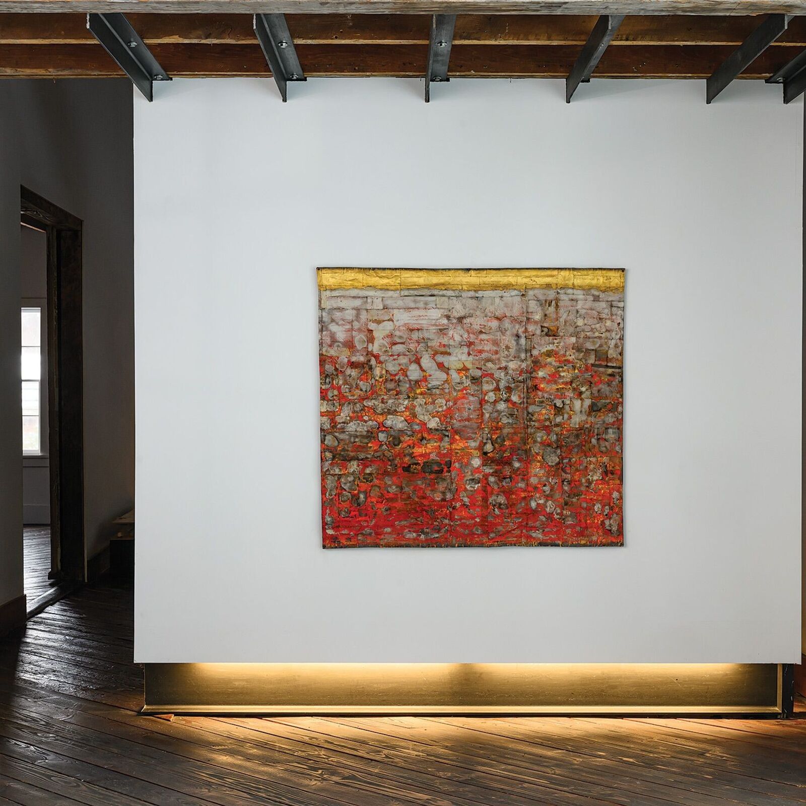 A textured abstract painting with red and gold tones hangs on a white wall in an industrial-style room.