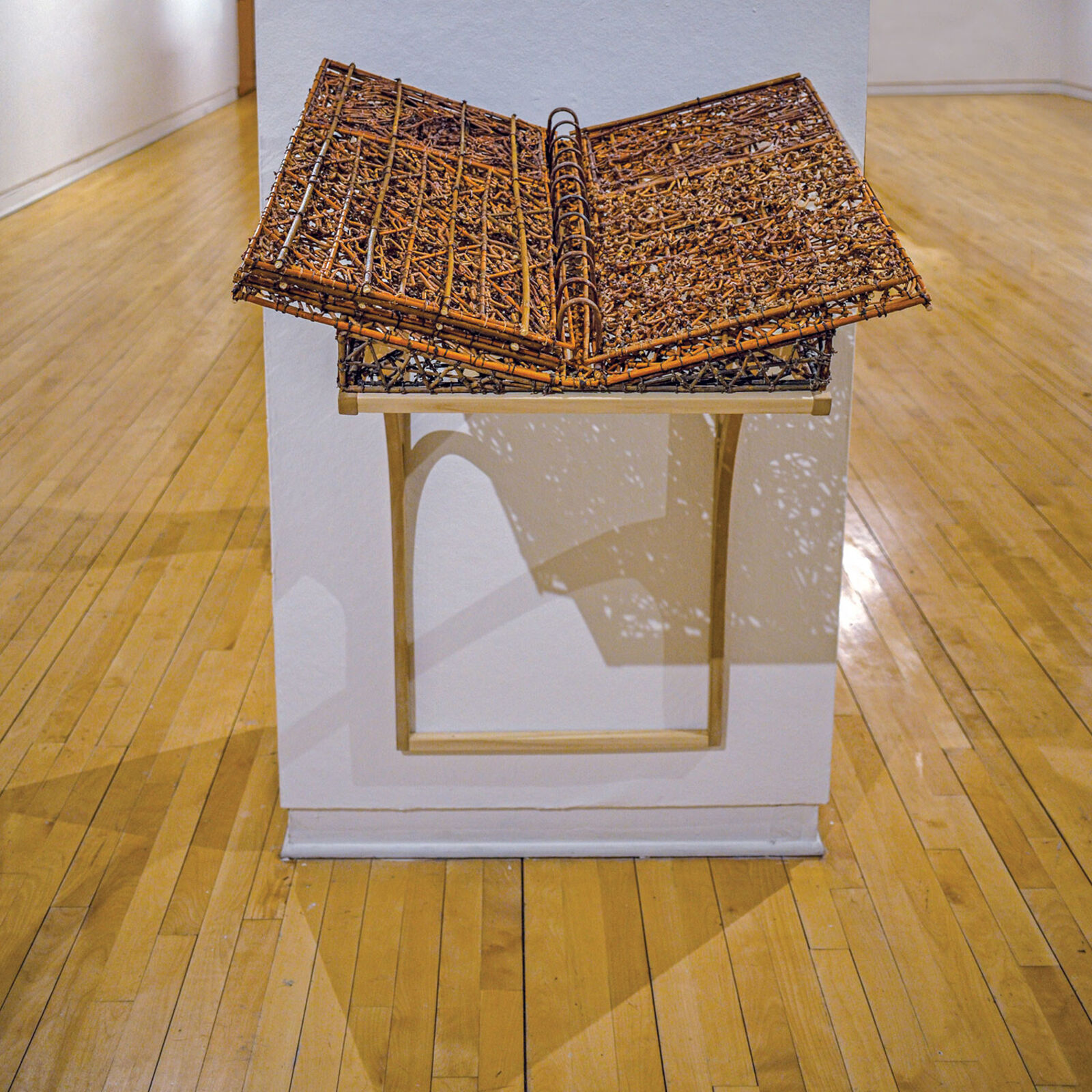 A sculptural book made of woven twigs and metal rings sits open on a white pedestal.