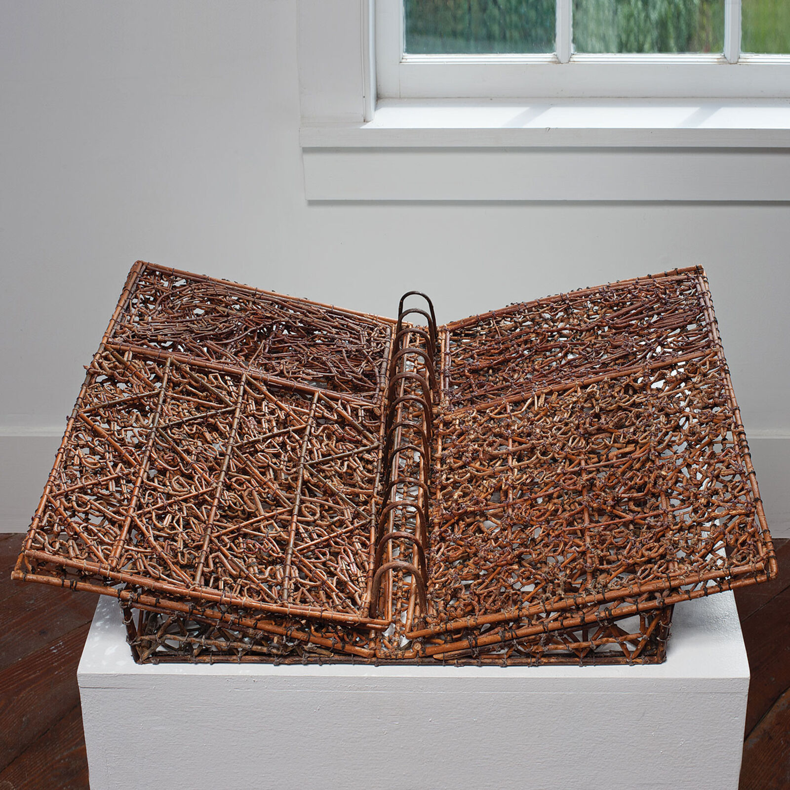 A sculptural book made of woven twigs and metal rings sits open on a white pedestal.