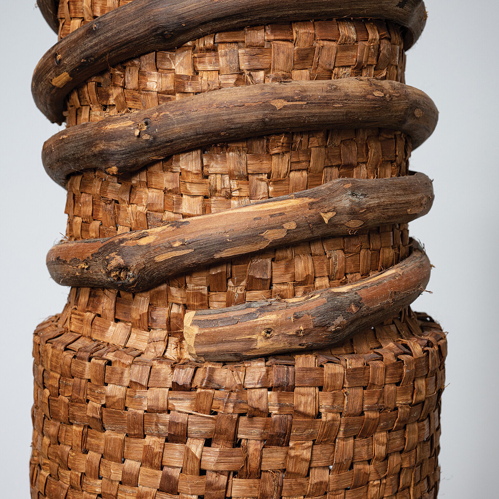 A tall woven basket sculpture wrapped with dark branches, displayed on a white pedestal.