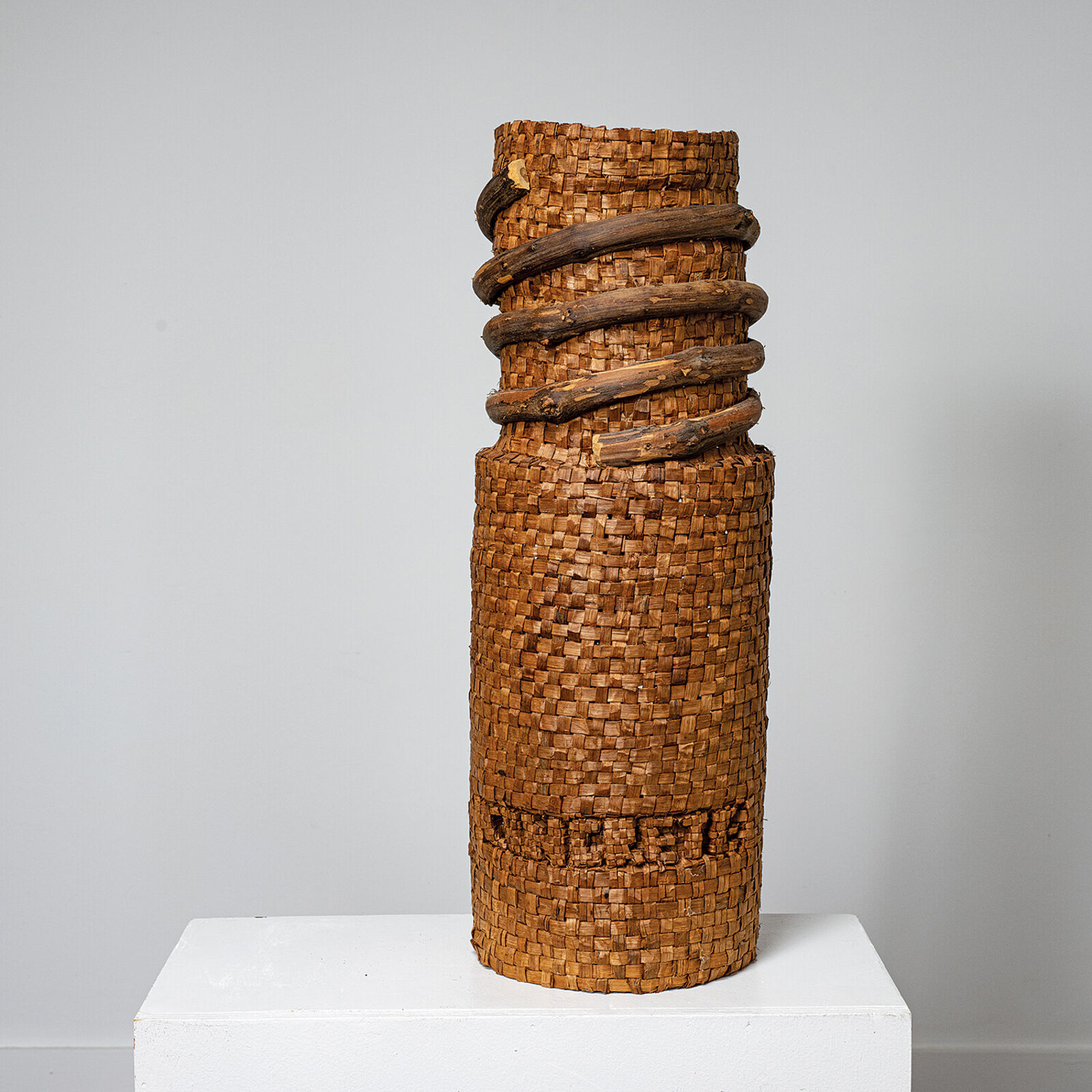 A tall woven basket sculpture wrapped with dark branches, displayed on a white pedestal.