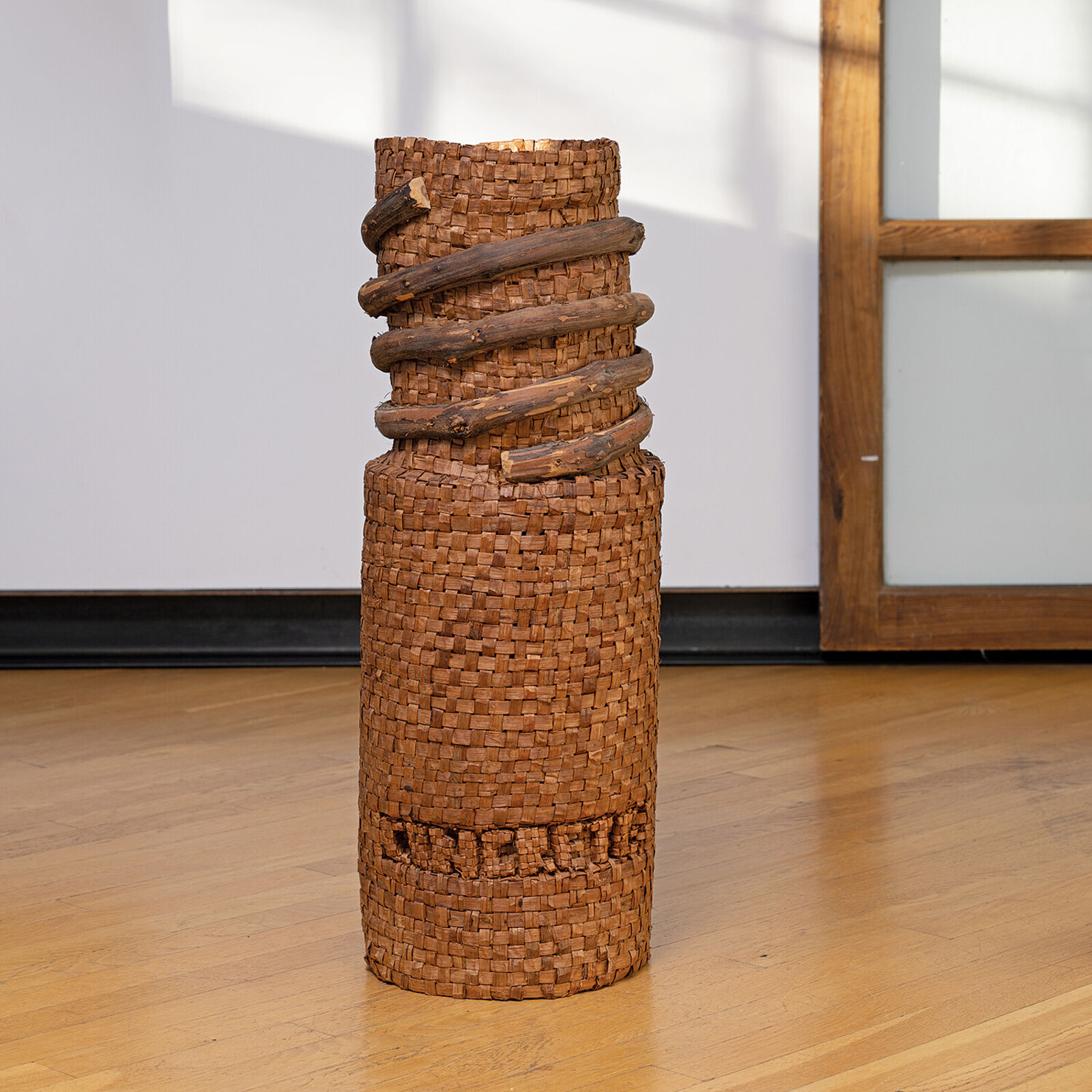 A tall woven basket sculpture wrapped with dark branches, displayed on a white pedestal.