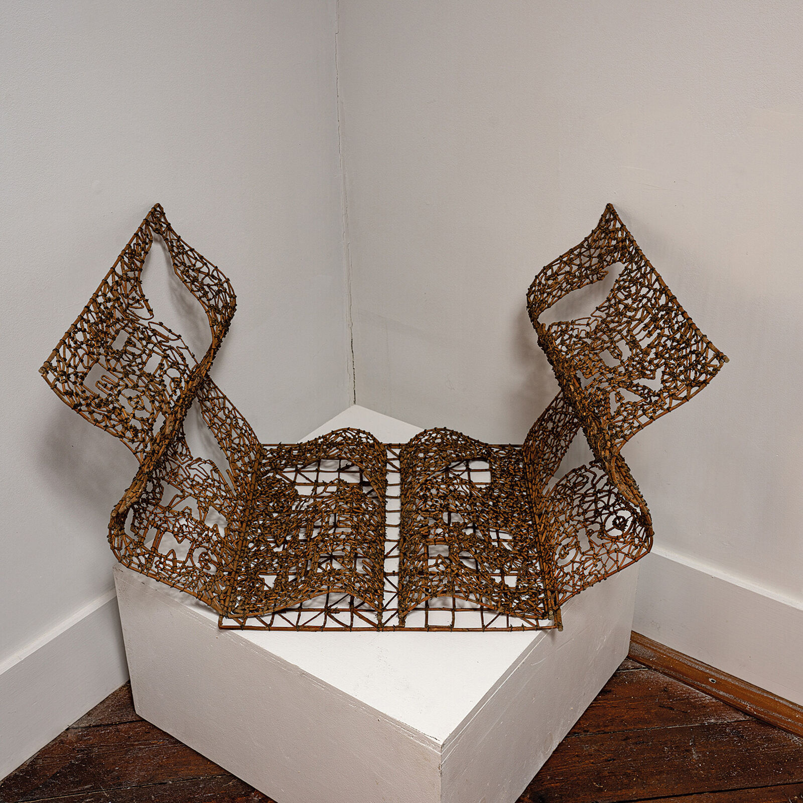 A sculptural book made of woven twigs sits on a white surface beneath a window.