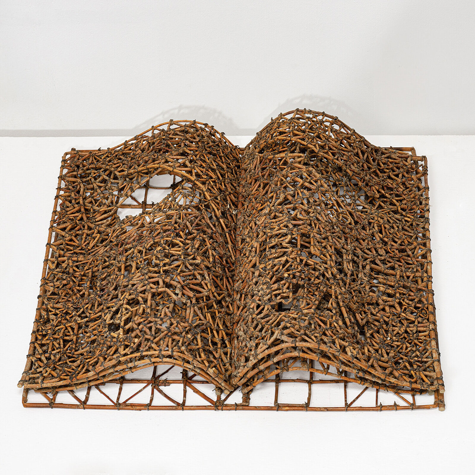 A sculptural book made of woven twigs sits on a white surface beneath a window.