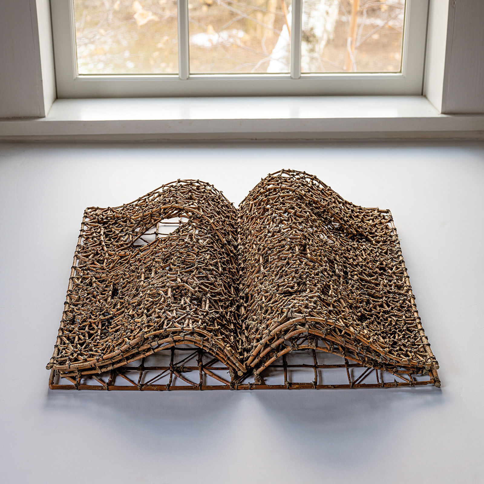 A sculptural book made of woven twigs sits on a white surface beneath a window.