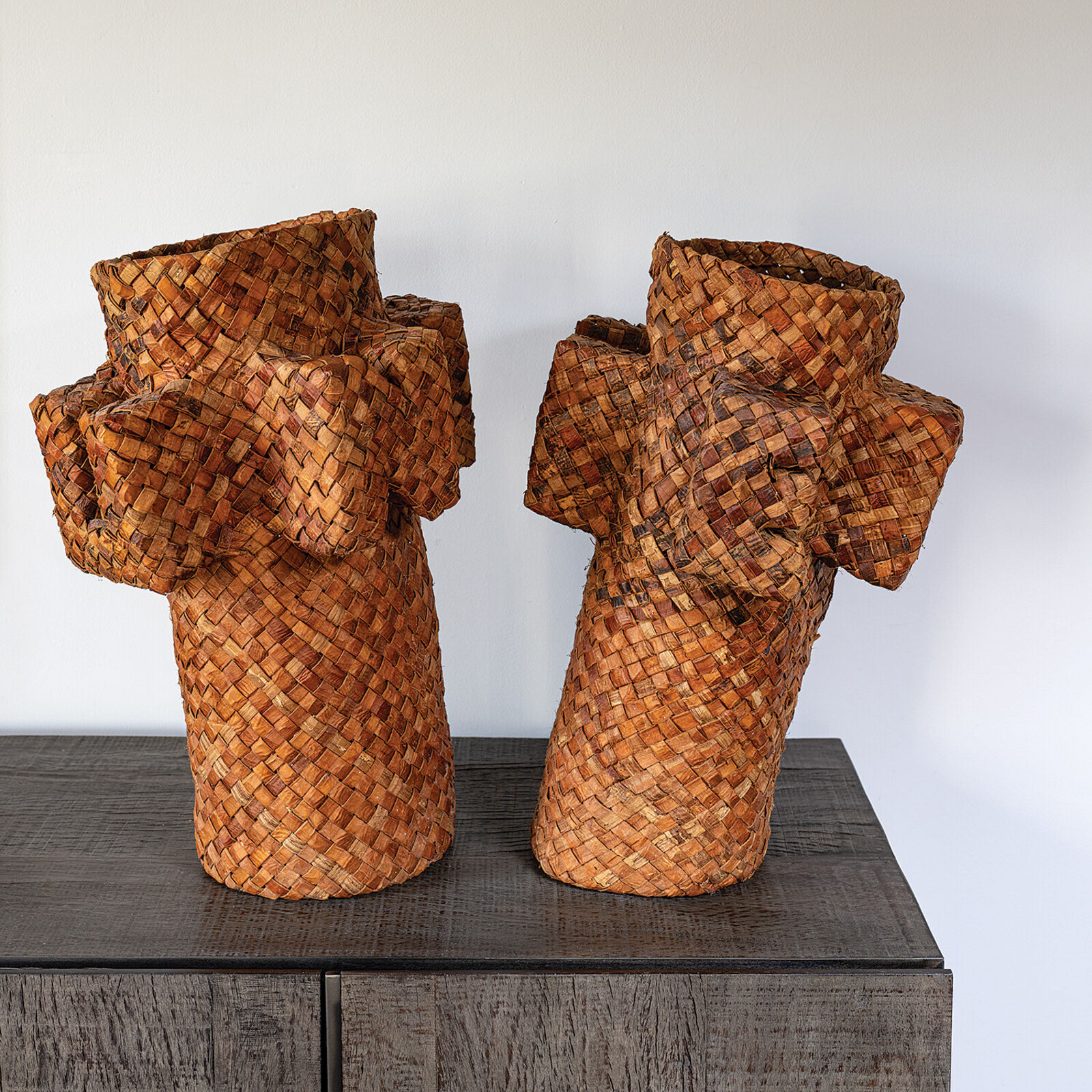 A pair of woven basket sculptures shaped like stylized figures with raised arms, displayed on a white pedestal.