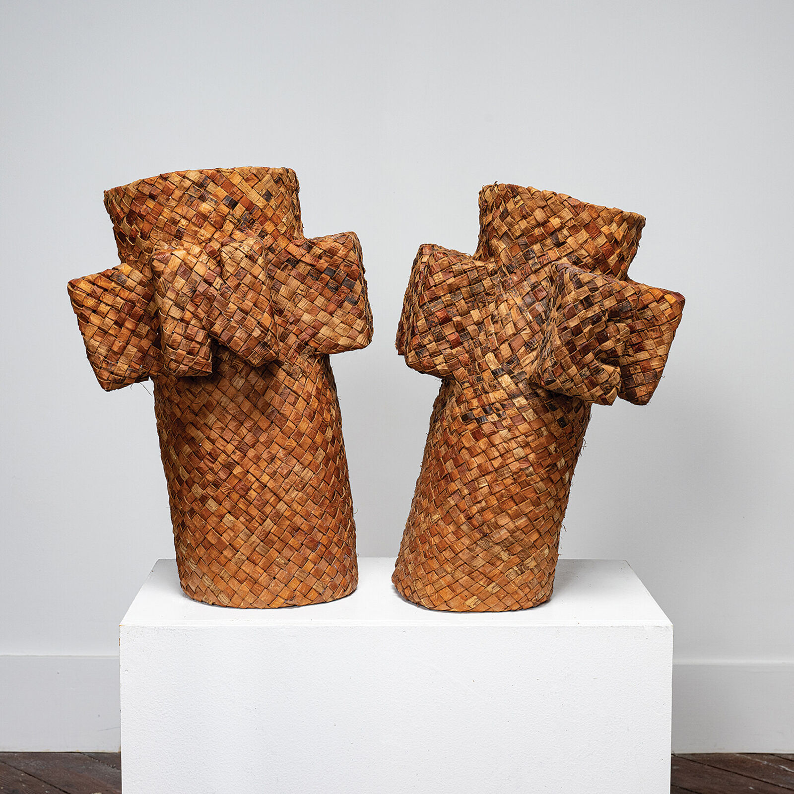 A pair of woven basket sculptures shaped like stylized figures with raised arms, displayed on a white pedestal.