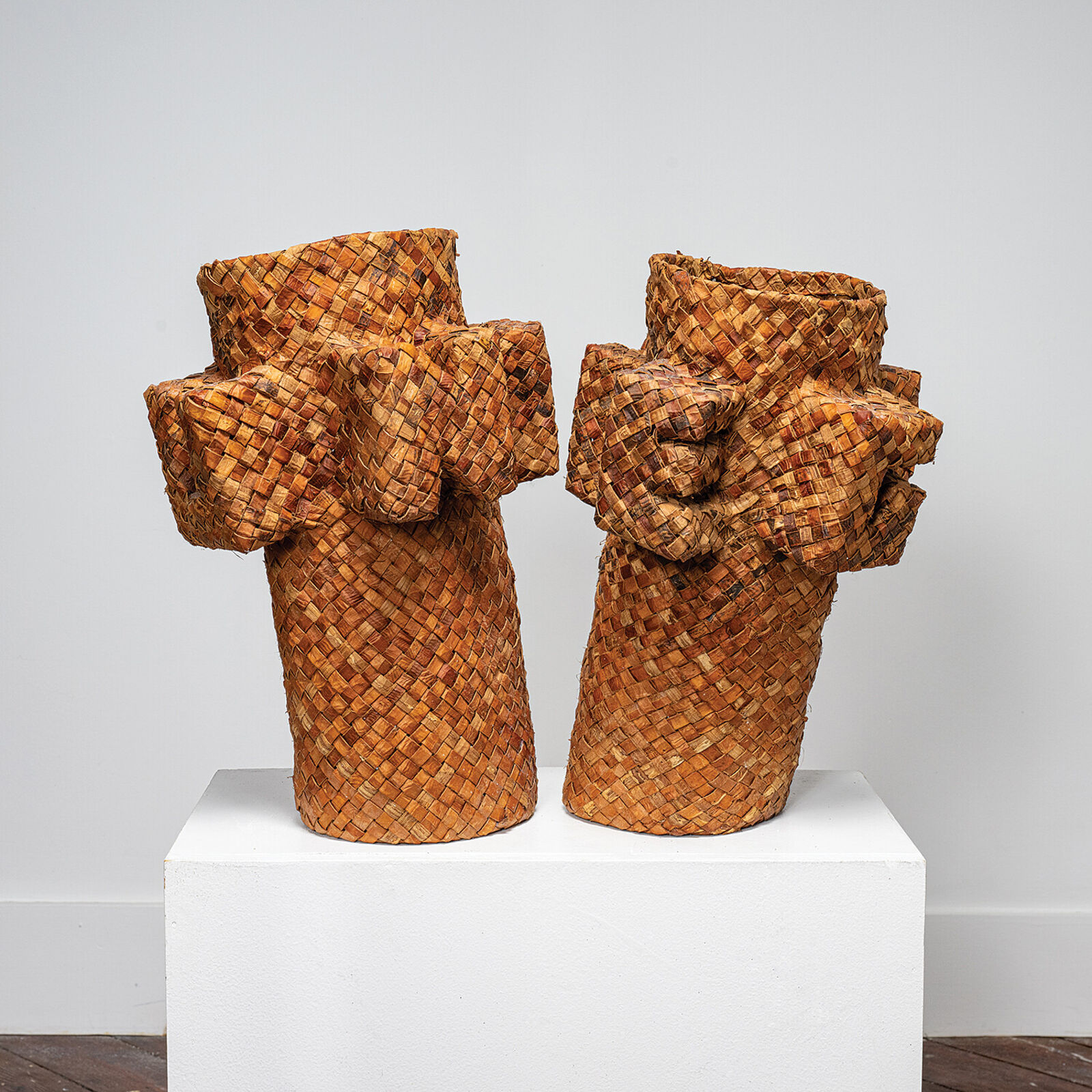 A pair of woven basket sculptures shaped like stylized figures with raised arms, displayed on a white pedestal.