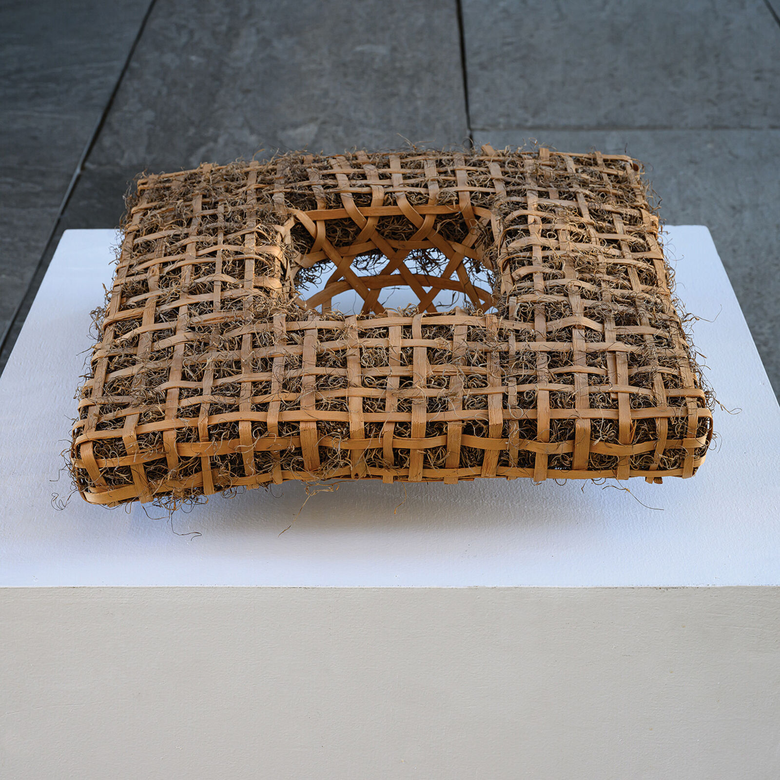 A woven rectangular sculpture with a central geometric cutout, displayed on a white pedestal against a grey tiled background.