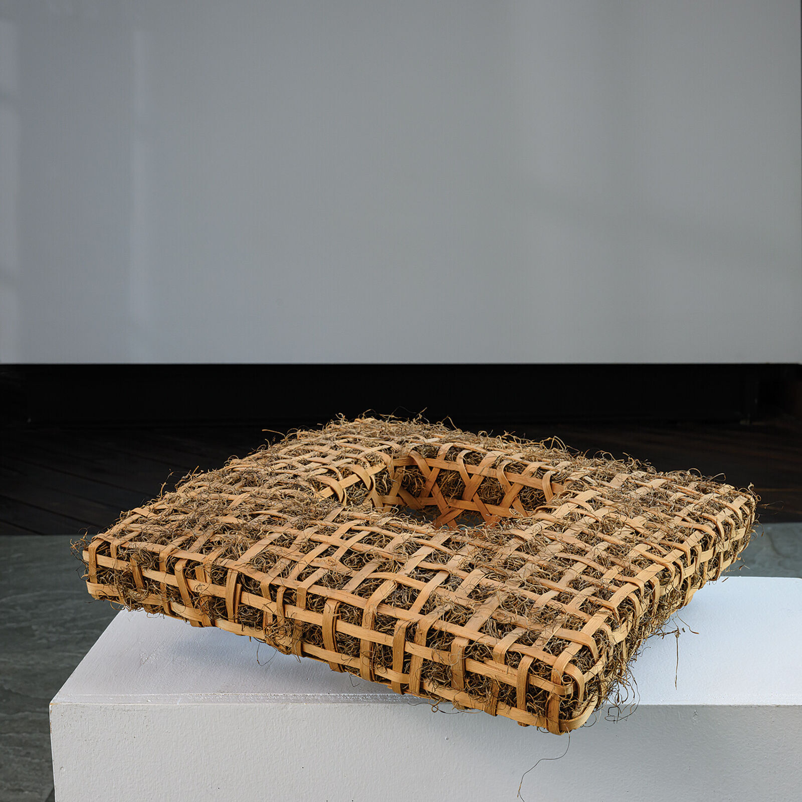 A woven rectangular sculpture with a central geometric cutout, displayed on a white pedestal against a grey tiled background.