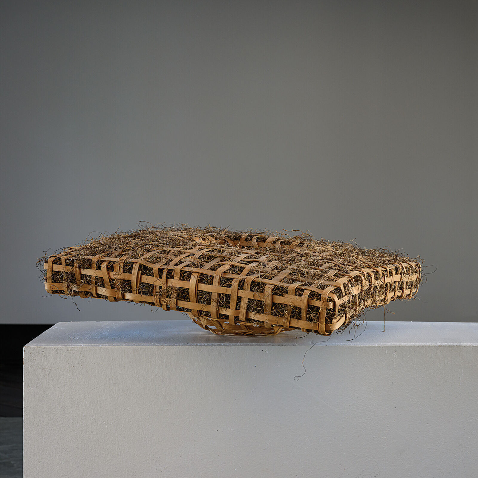 A woven rectangular sculpture with a central geometric cutout, displayed on a white pedestal against a grey tiled background.