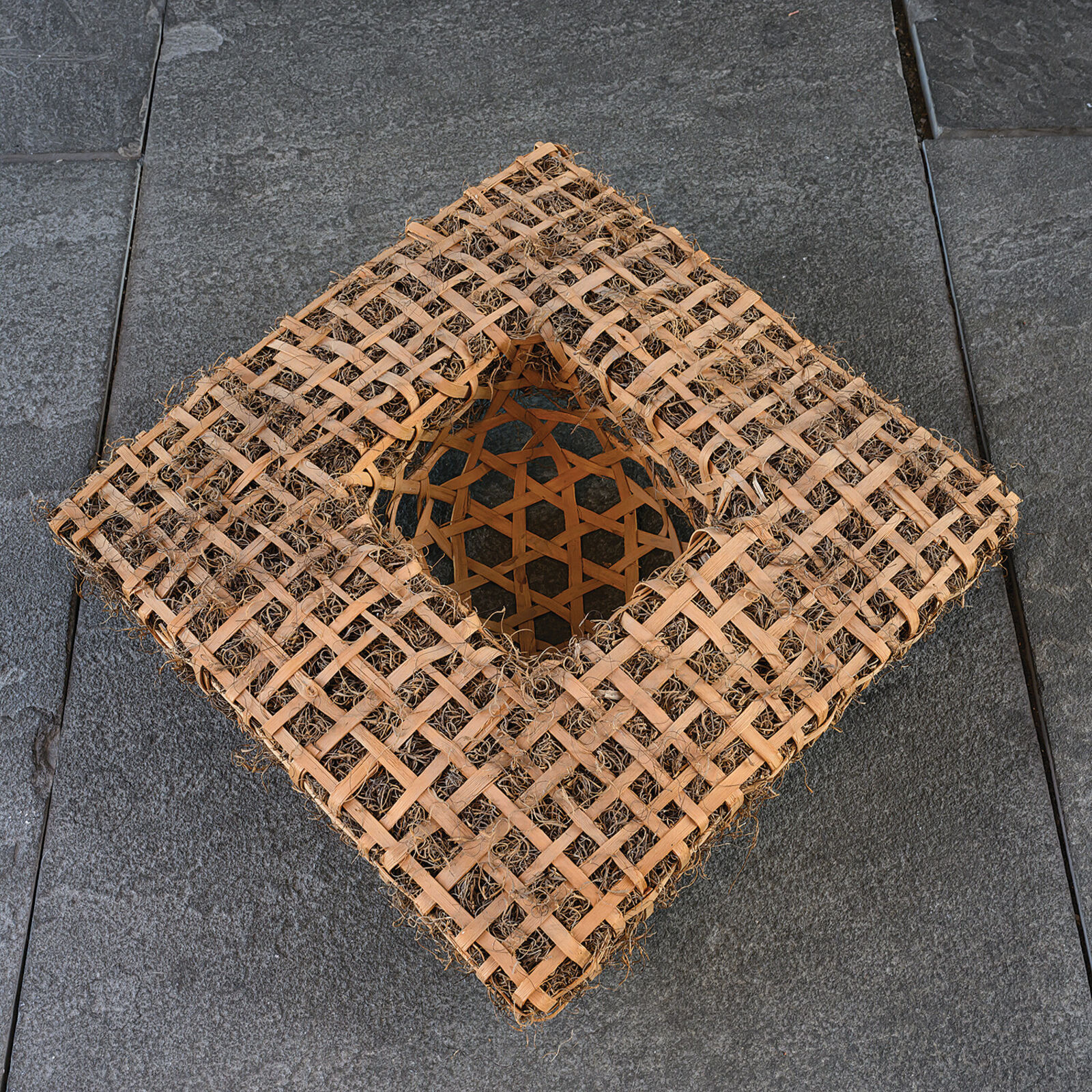 A woven rectangular sculpture with a central geometric cutout, displayed on a white pedestal against a grey tiled background.