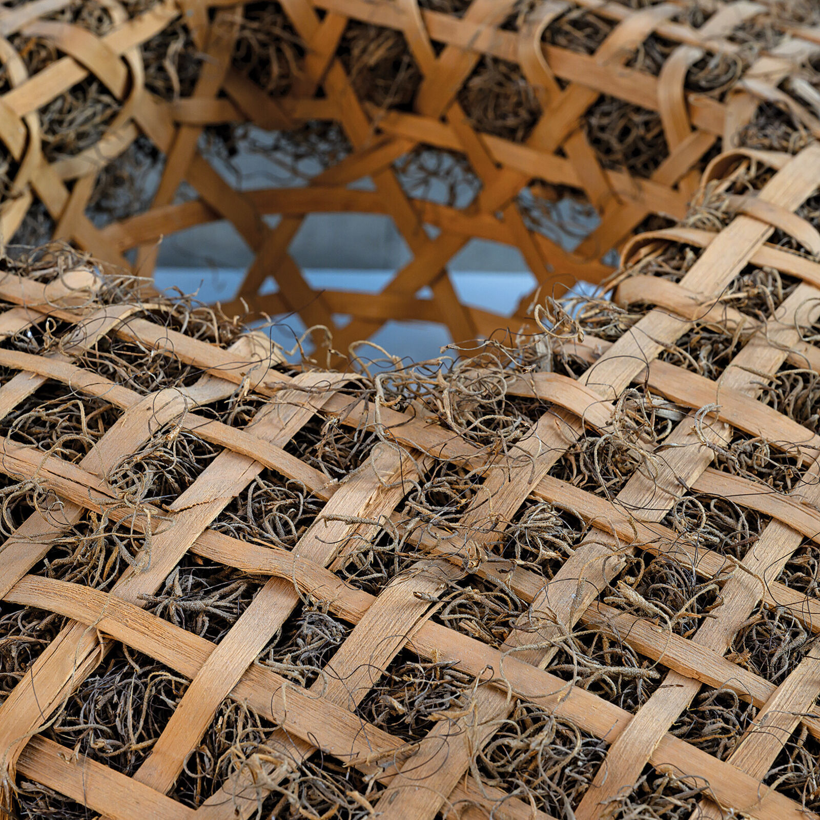 A woven rectangular sculpture with a central geometric cutout, displayed on a white pedestal against a grey tiled background.