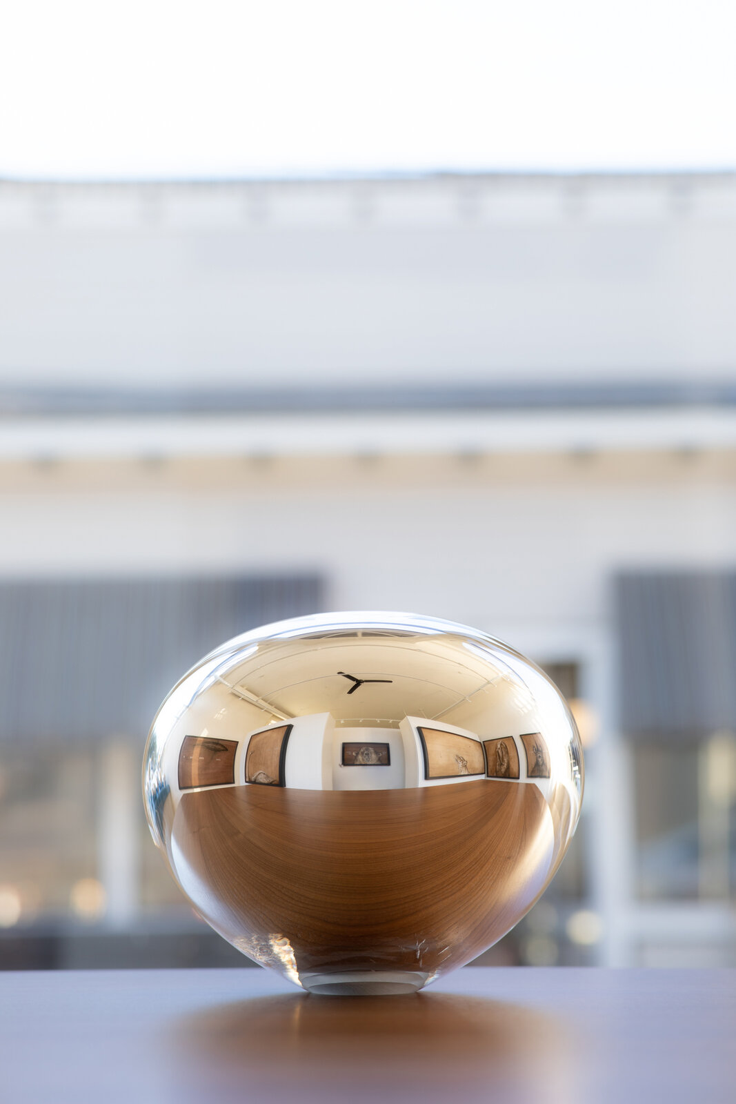 A polished wooden sphere reflecting a gallery interior, set against a blurred architectural backdrop.