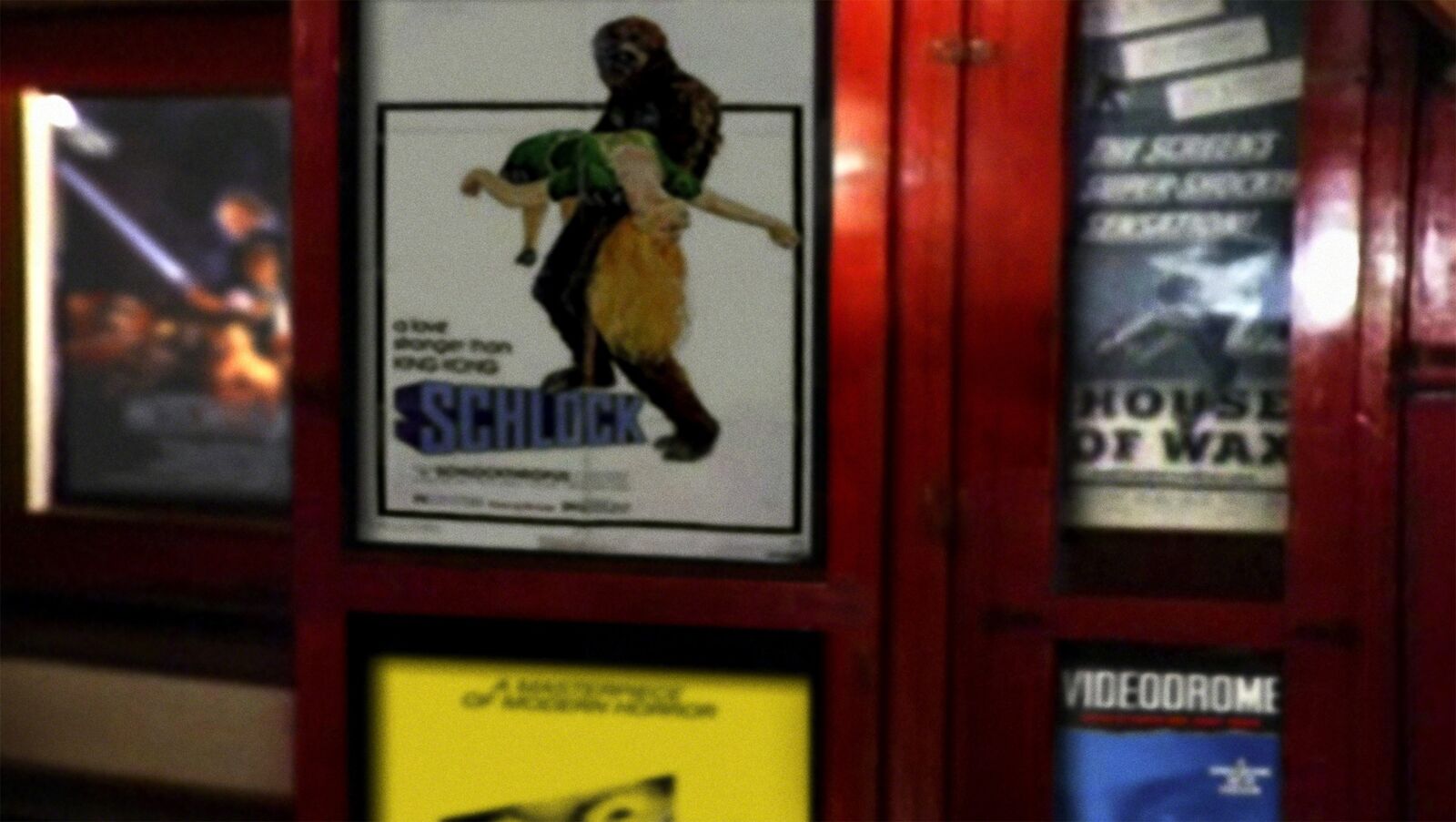 A dimly lit poster wall featuring a central framed artwork of two figures in dynamic motion, surrounded by other posters and red wooden frames.
