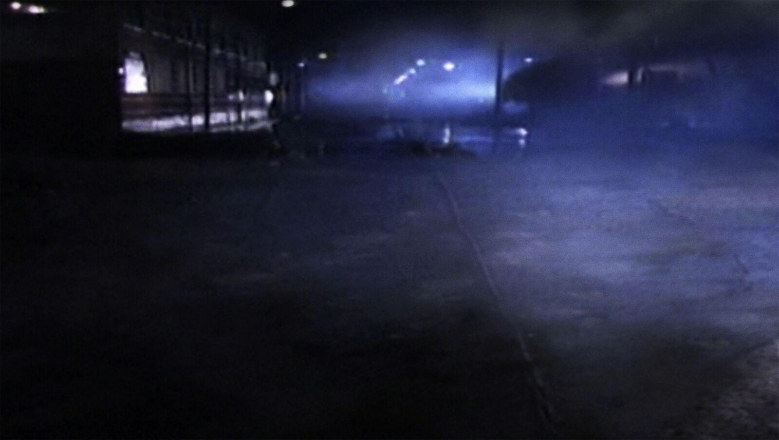 A dark, atmospheric night scene with a blue-lit foggy street and distant building lights.