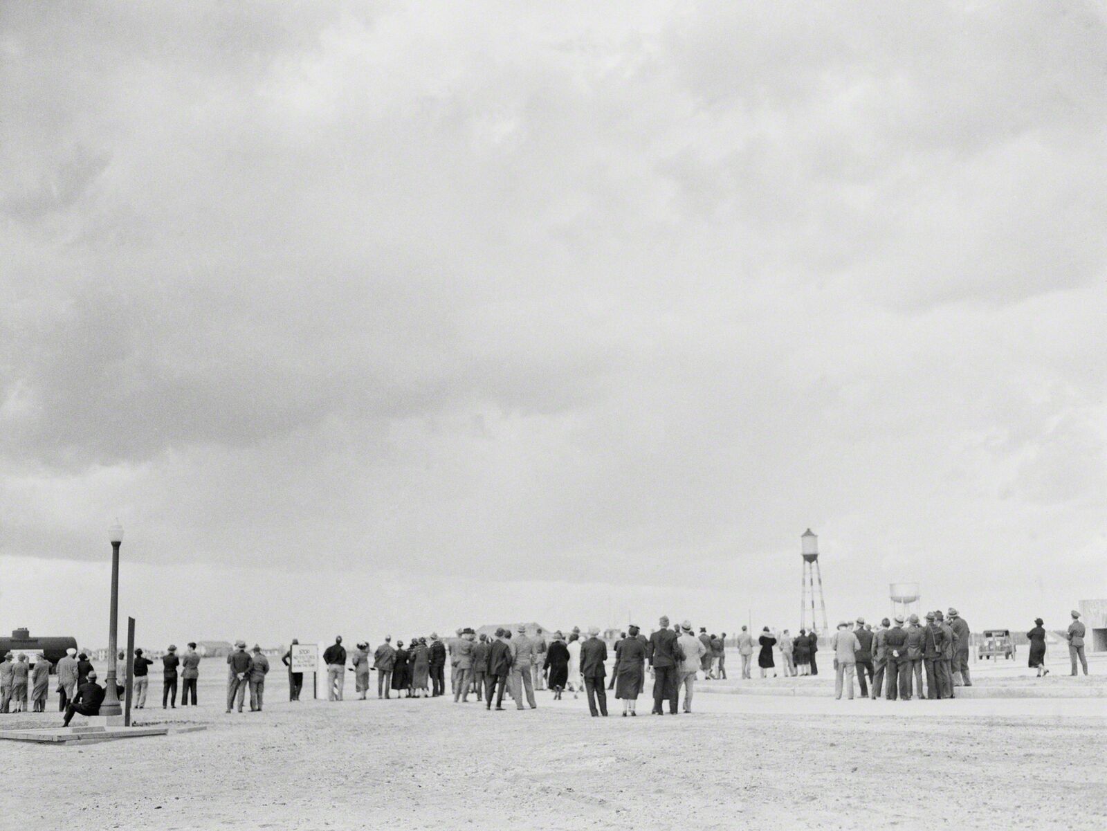 A black and white photograph captures a large gathering of people in an open, barren landscape under a heavy sky.
