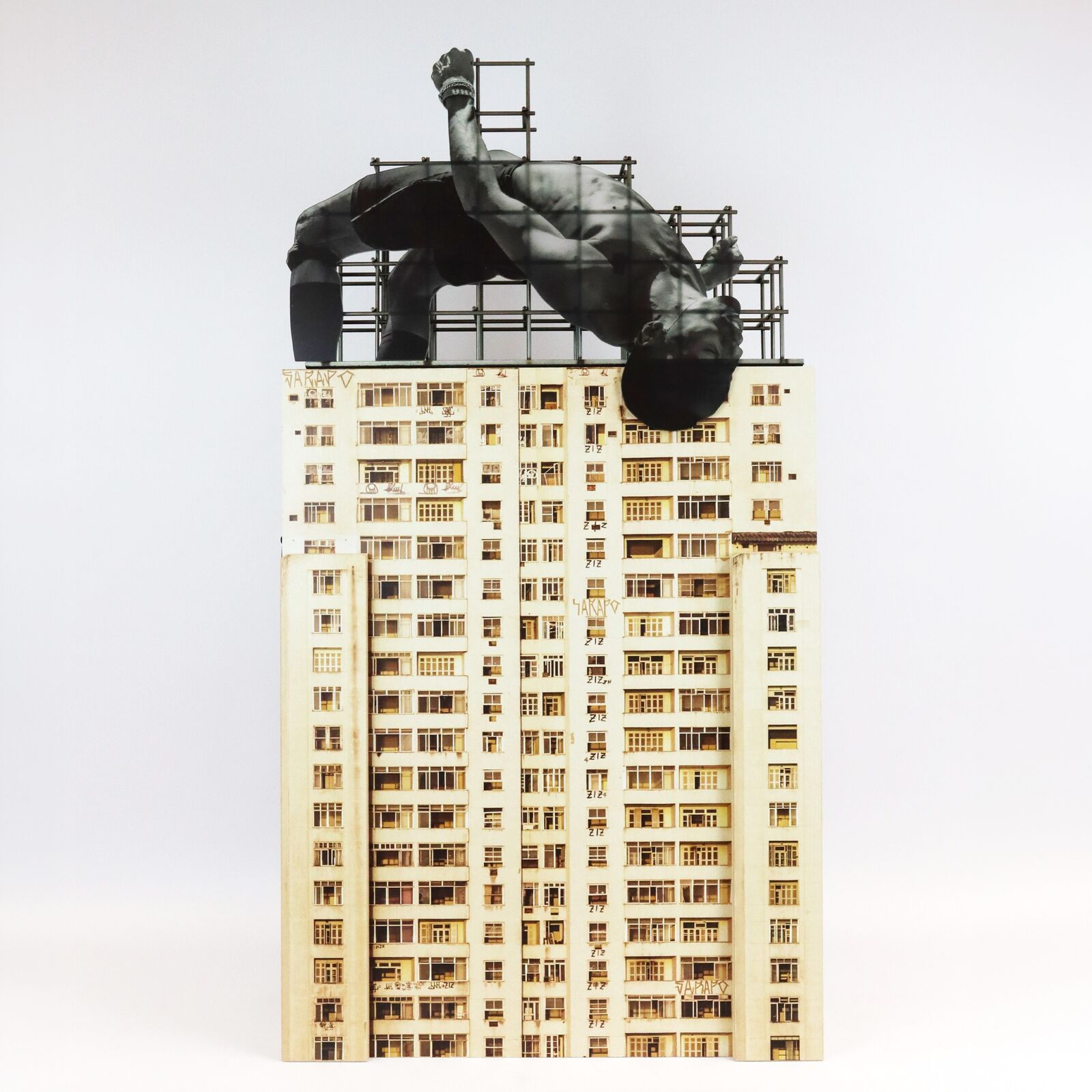 A miniature architectural model of a high-rise building topped by a suspended, contorted human figure.