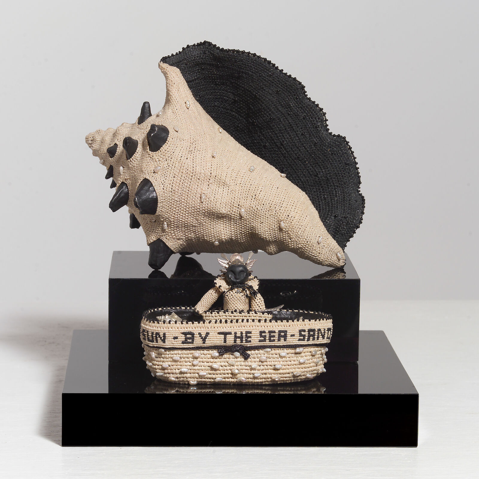 A sculptural arrangement featuring a large beaded shell resting on a black block above a woven basket with text.