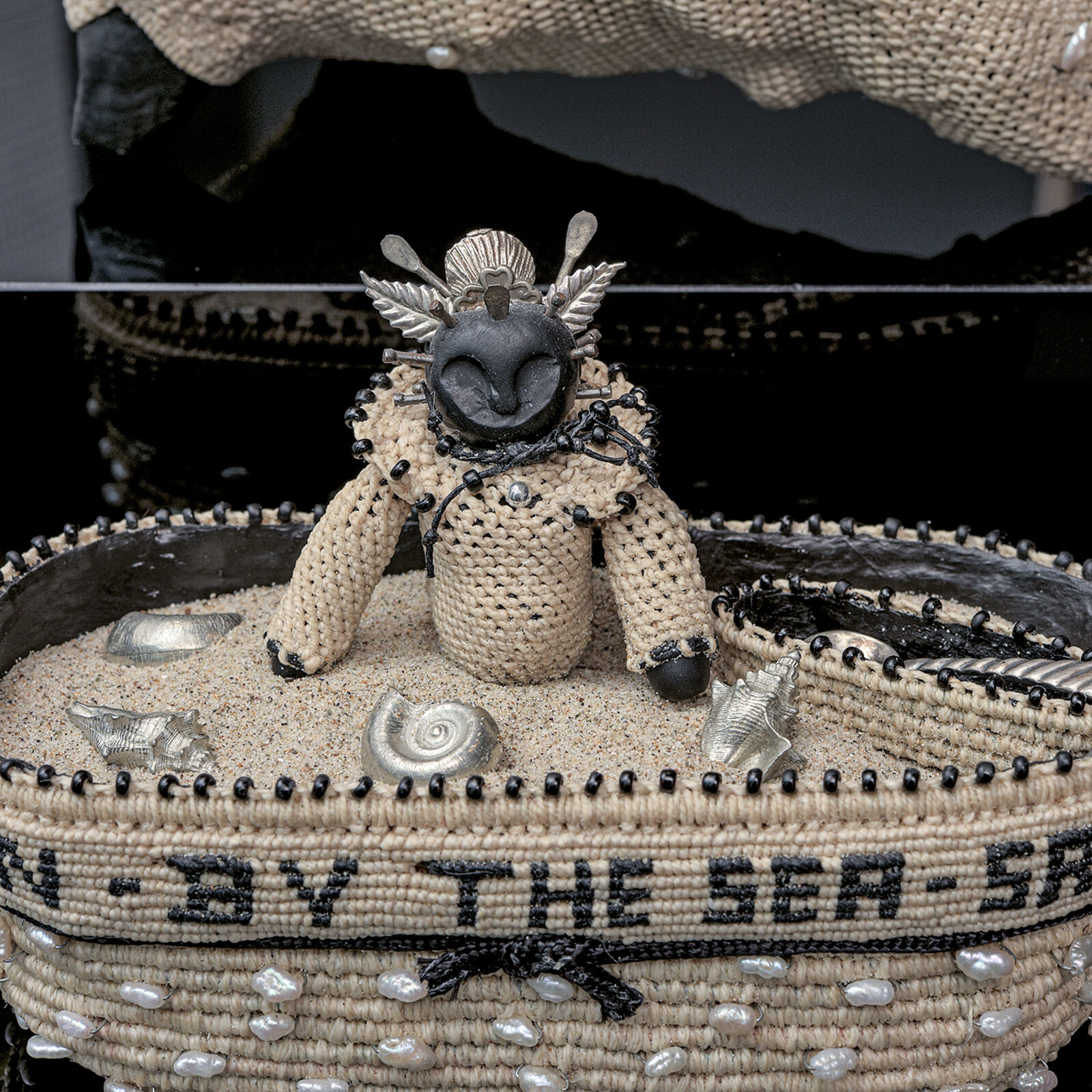 A sculptural arrangement featuring a large beaded shell resting on a black block above a woven basket with text.