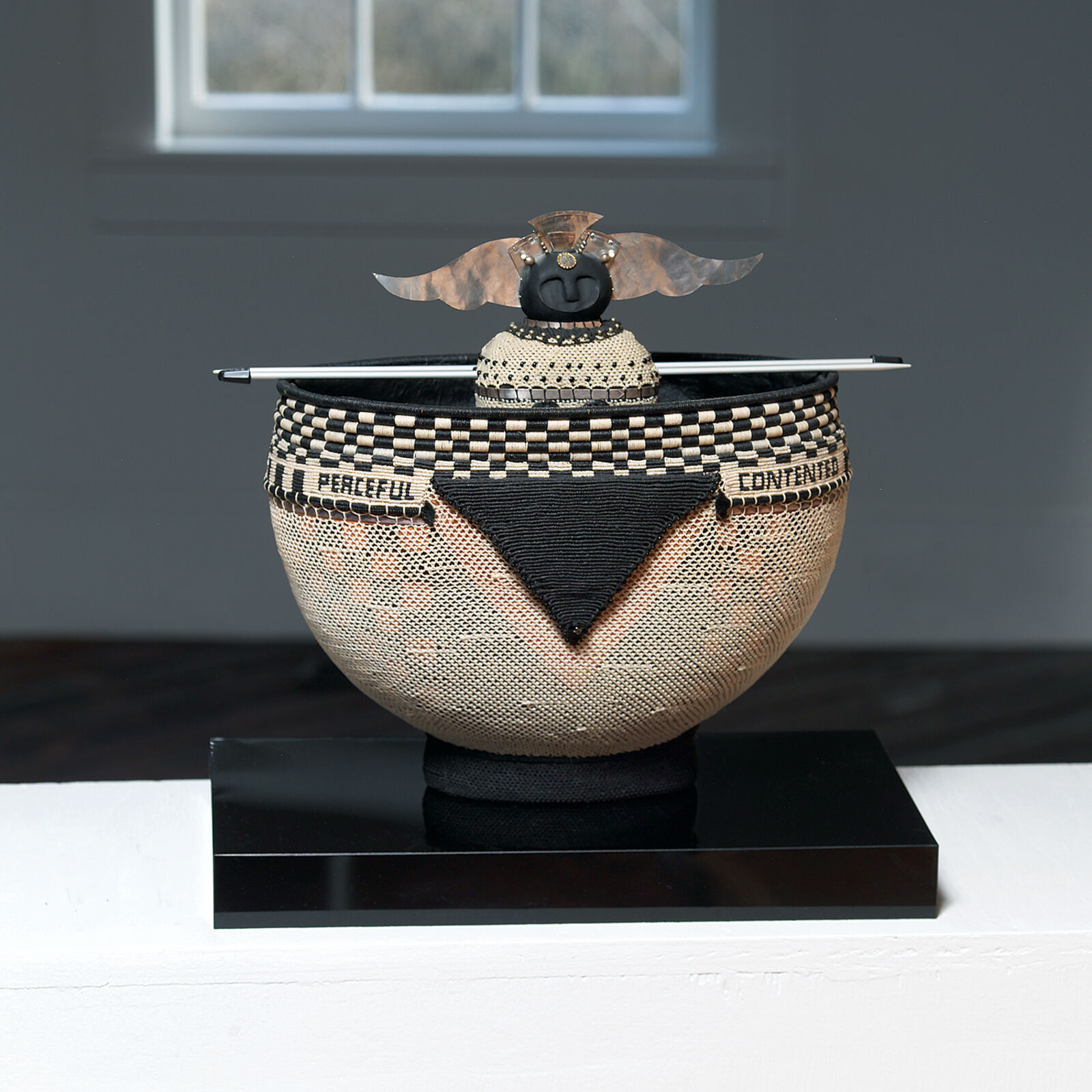 A woven bowl with a sculptural lid featuring a face and wings, resting on a black base.