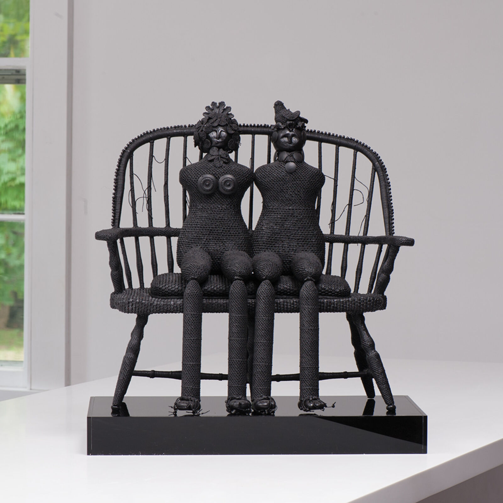 A monochromatic sculpture of two figures seated on a bench, crafted from textured black material.