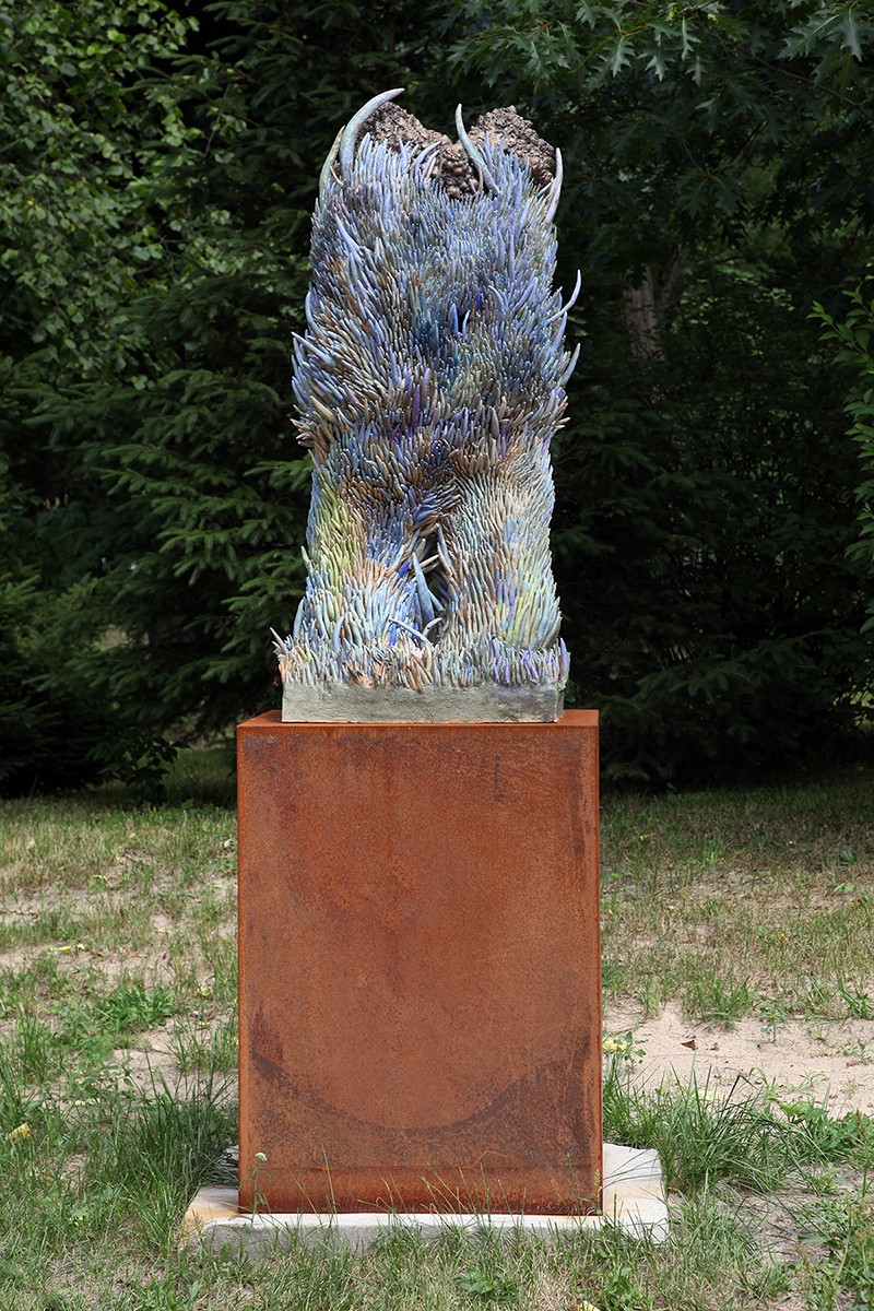 A sculpture of a bird sitting on top of a metal pedestal in the grass.