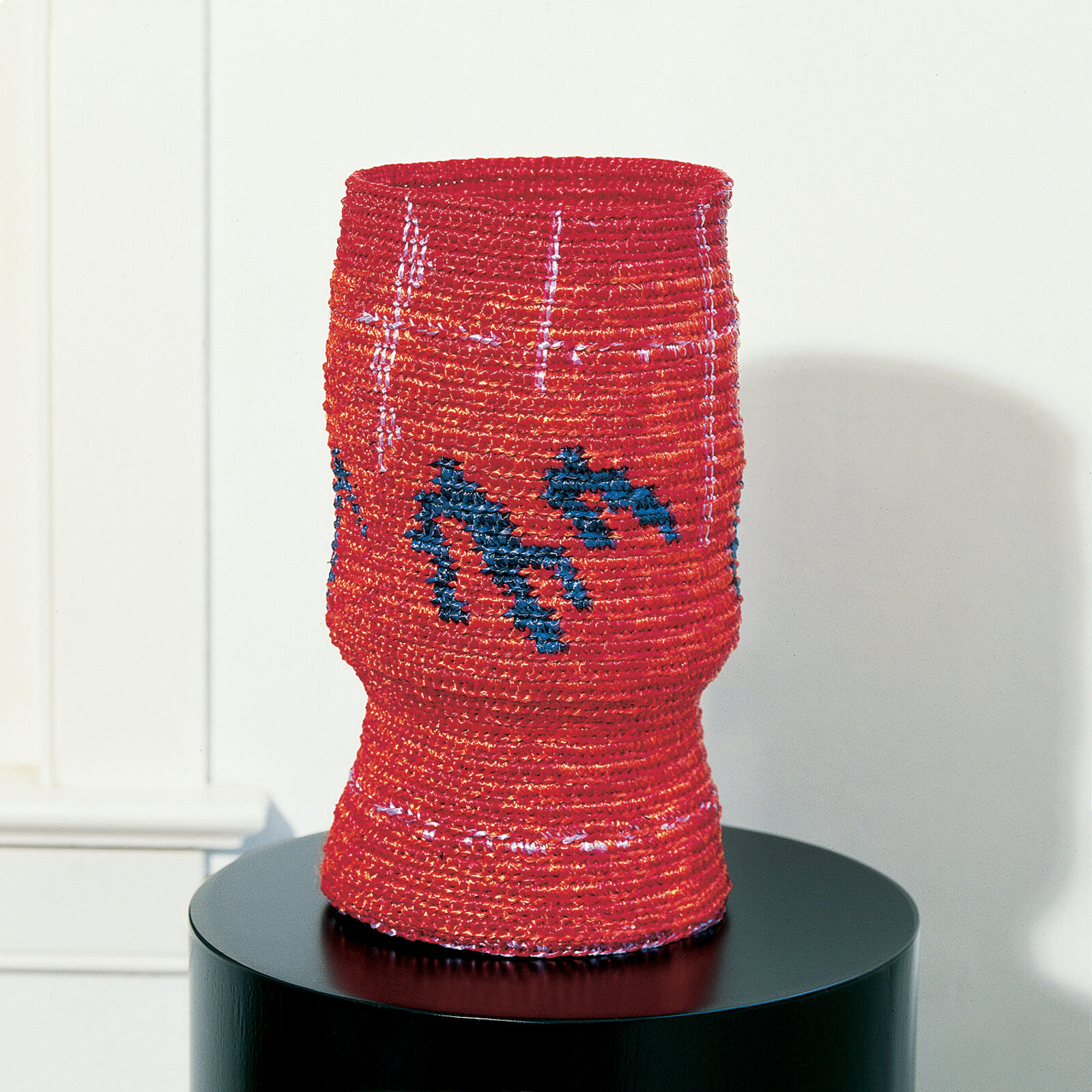 A red beaded vase with blue geometric patterns displayed on a black pedestal.