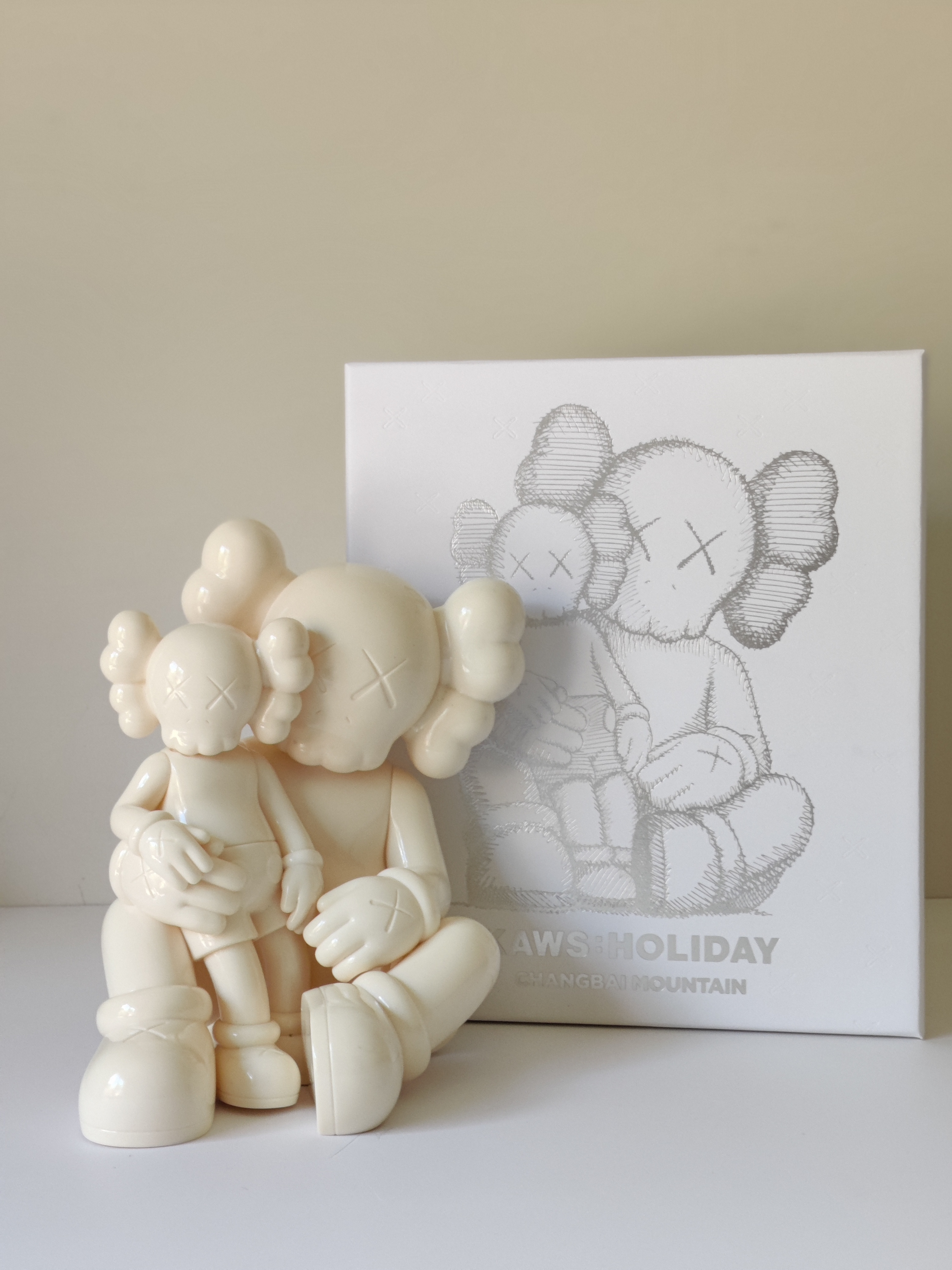 A glossy white ceramic sculpture of two stylized figures with cloud-like hair and X-shaped eyes, one seated and holding the other.