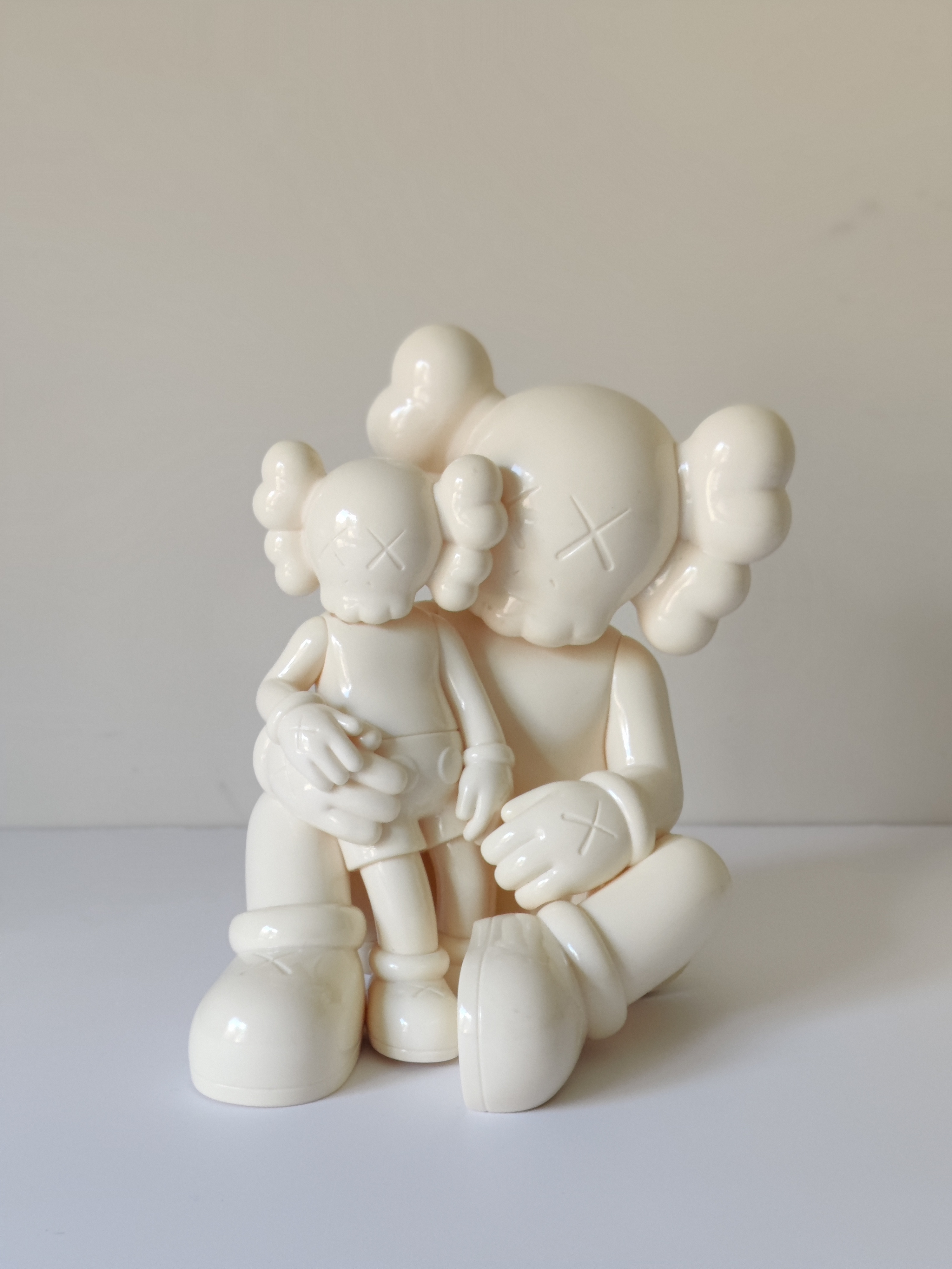 A glossy white ceramic sculpture of two stylized figures with cloud-like hair and X-shaped eyes, one seated and holding the other.