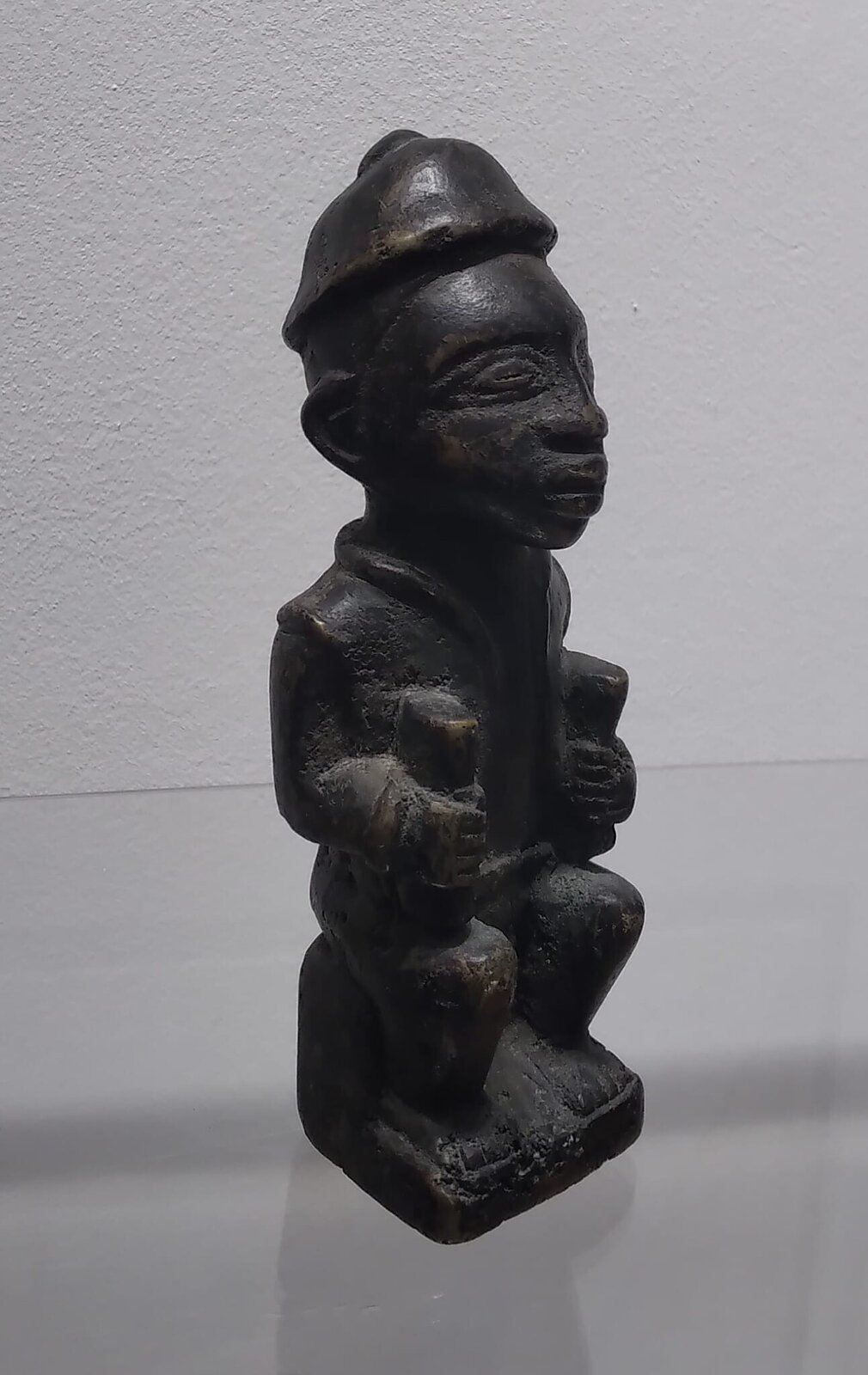 A dark, squat sculpture of a seated figure in profile against a plain background.