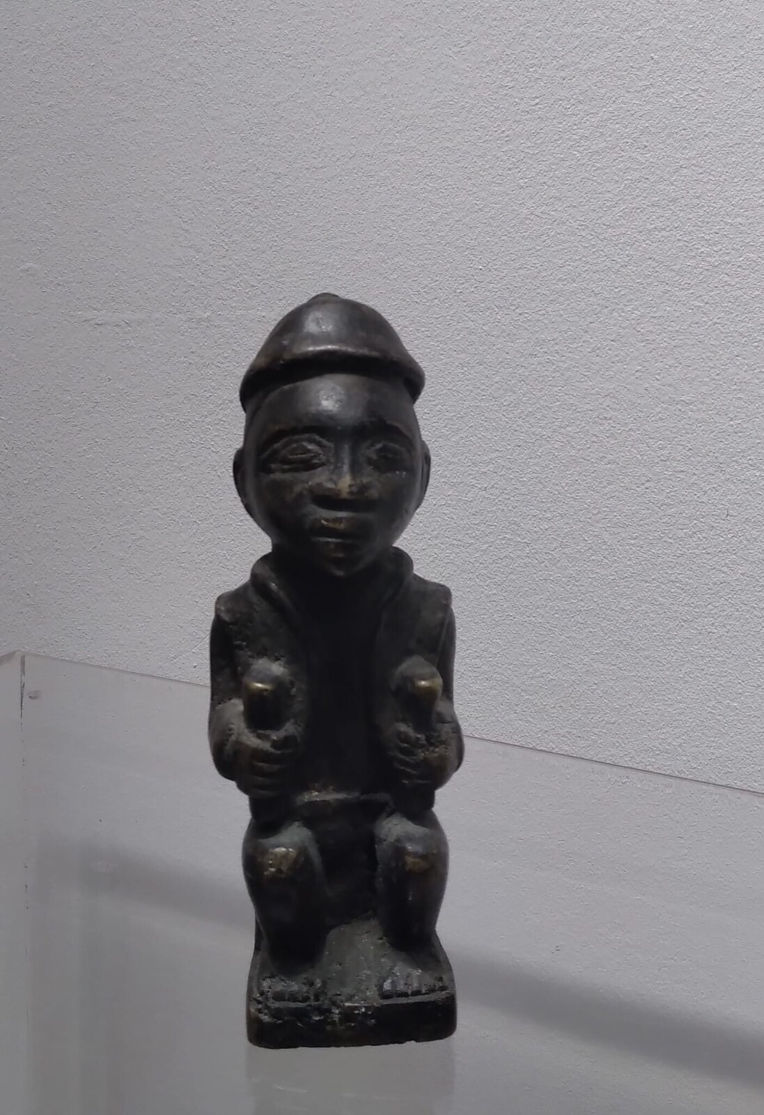 A dark, squat sculpture of a seated figure in profile against a plain background.