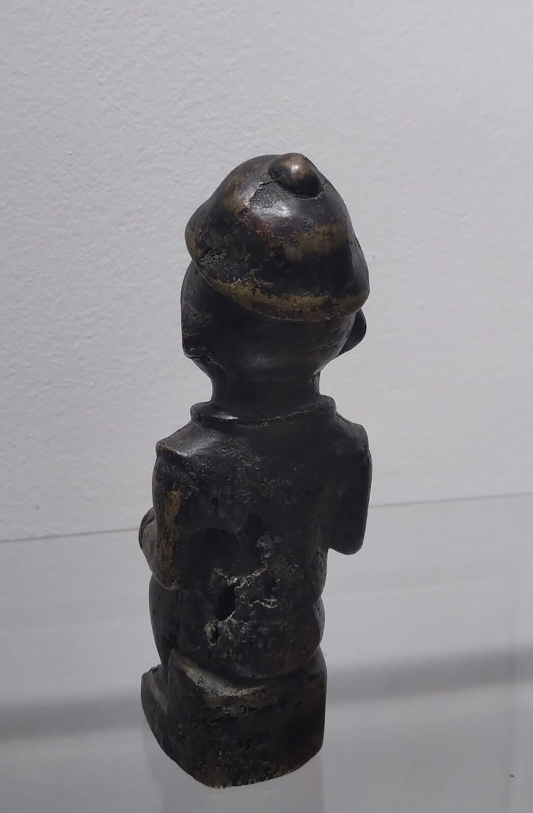 A dark, squat sculpture of a seated figure in profile against a plain background.