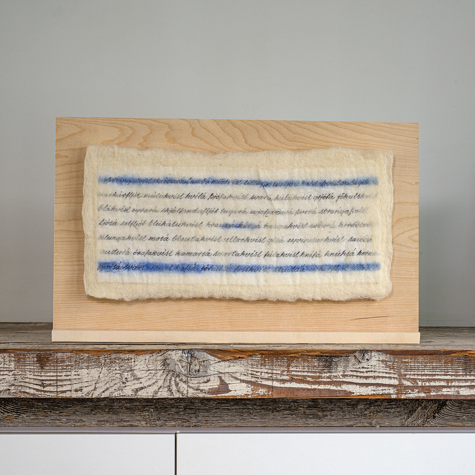 A minimalist textile artwork featuring blue and black script on a cream-colored felt, mounted on light wood.