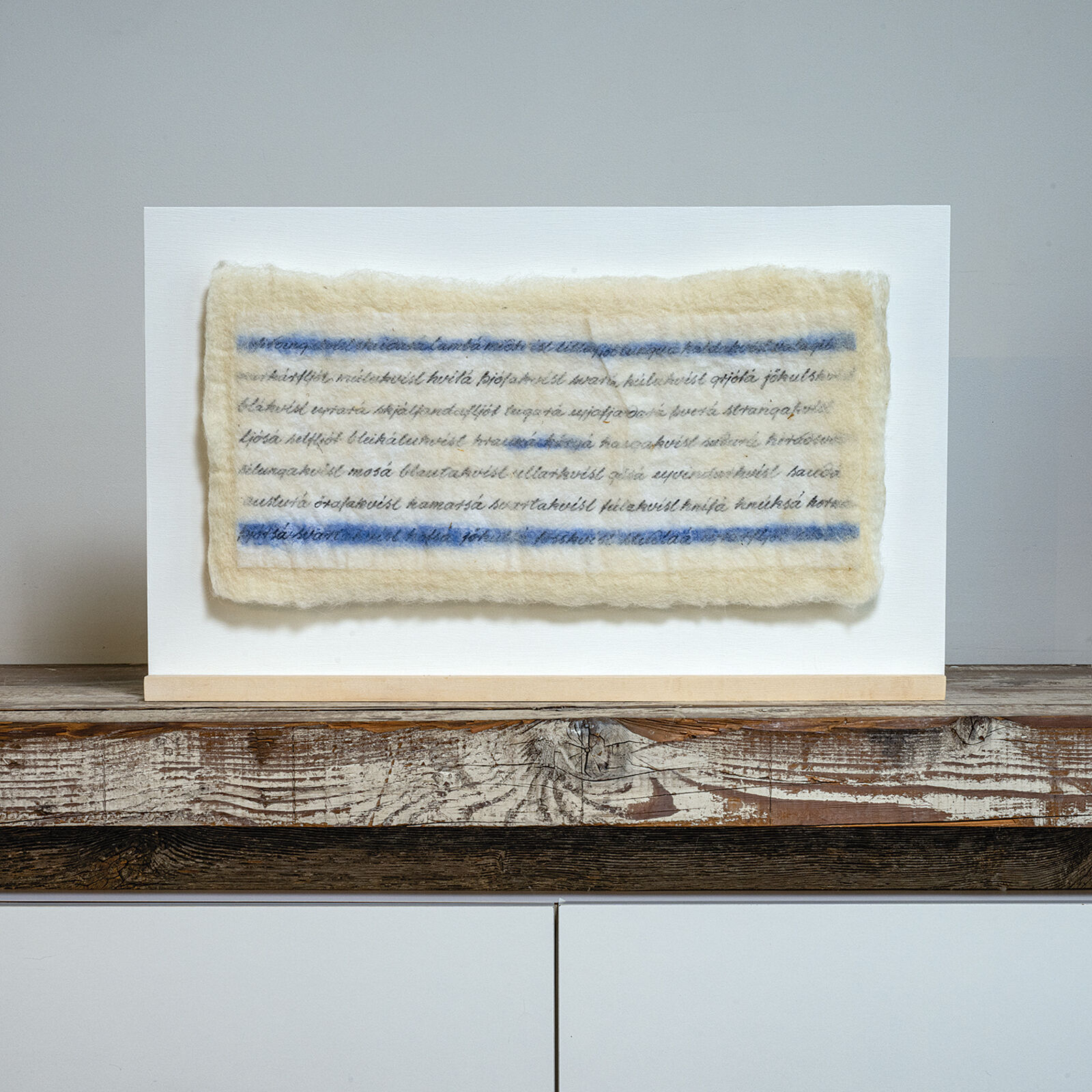 A minimalist textile artwork featuring blue and black script on a cream-colored felt, mounted on light wood.
