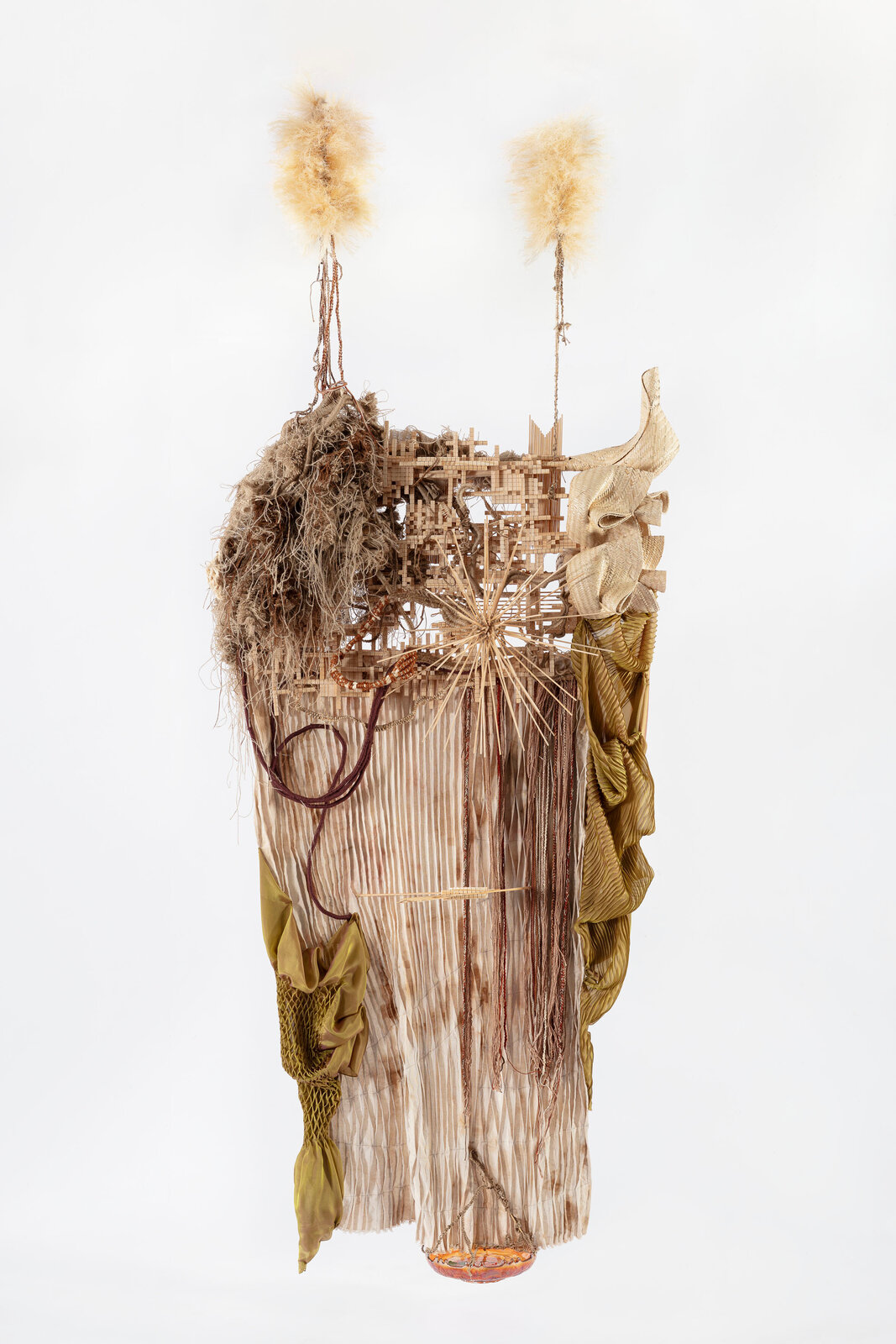 A complex mixed-media sculpture featuring natural fibers, woven elements, and hanging organic forms against a stark white background.