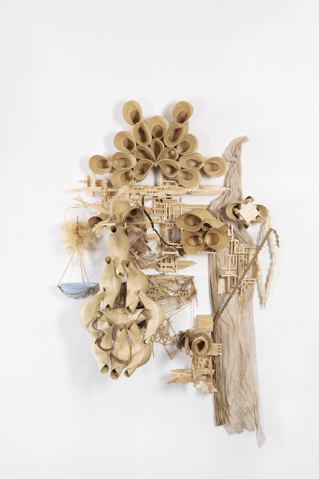 A complex mixed-media sculpture composed of straw, wood, and fabric elements arranged in an abstract composition.