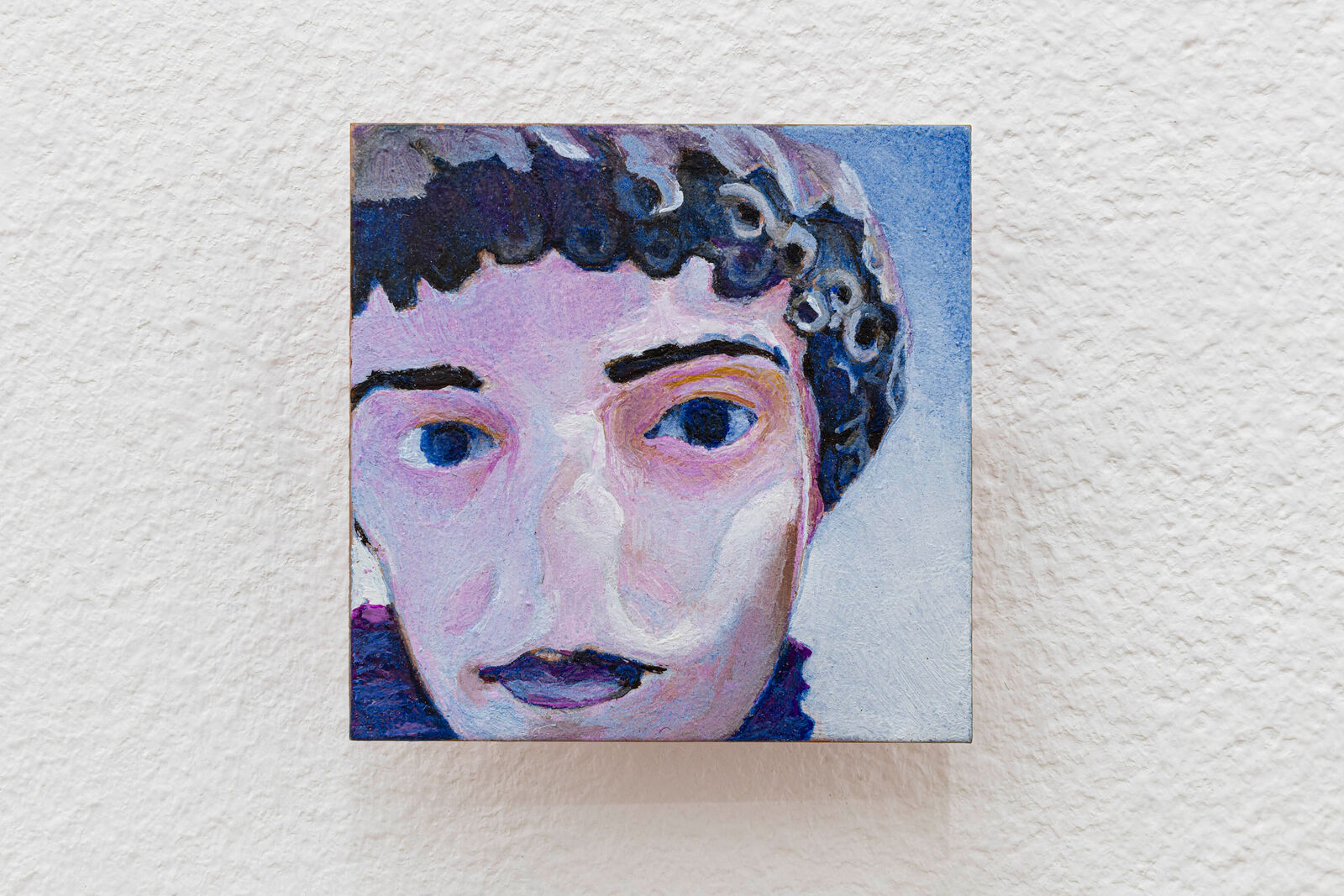 A small oil painting of a face with curly hair, rendered in bold colors against a textured white wall.