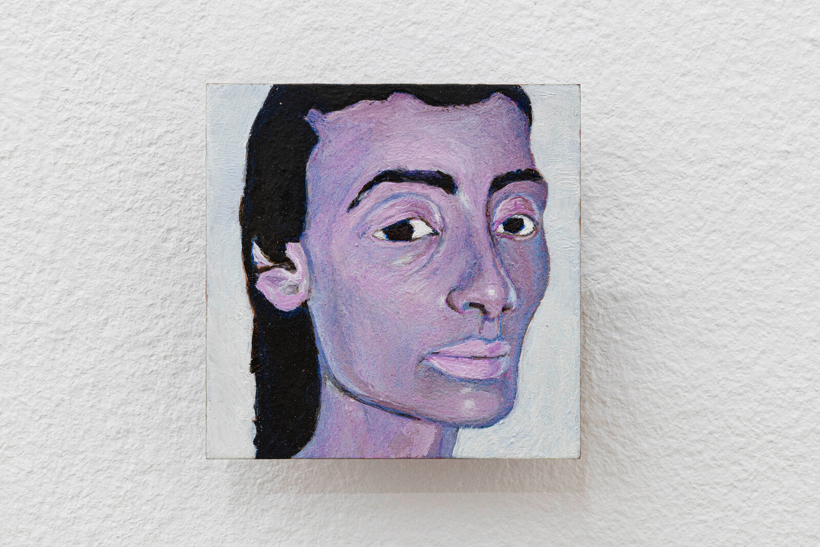 A small oil painting of a face with purple skin and black hair against a white textured wall.